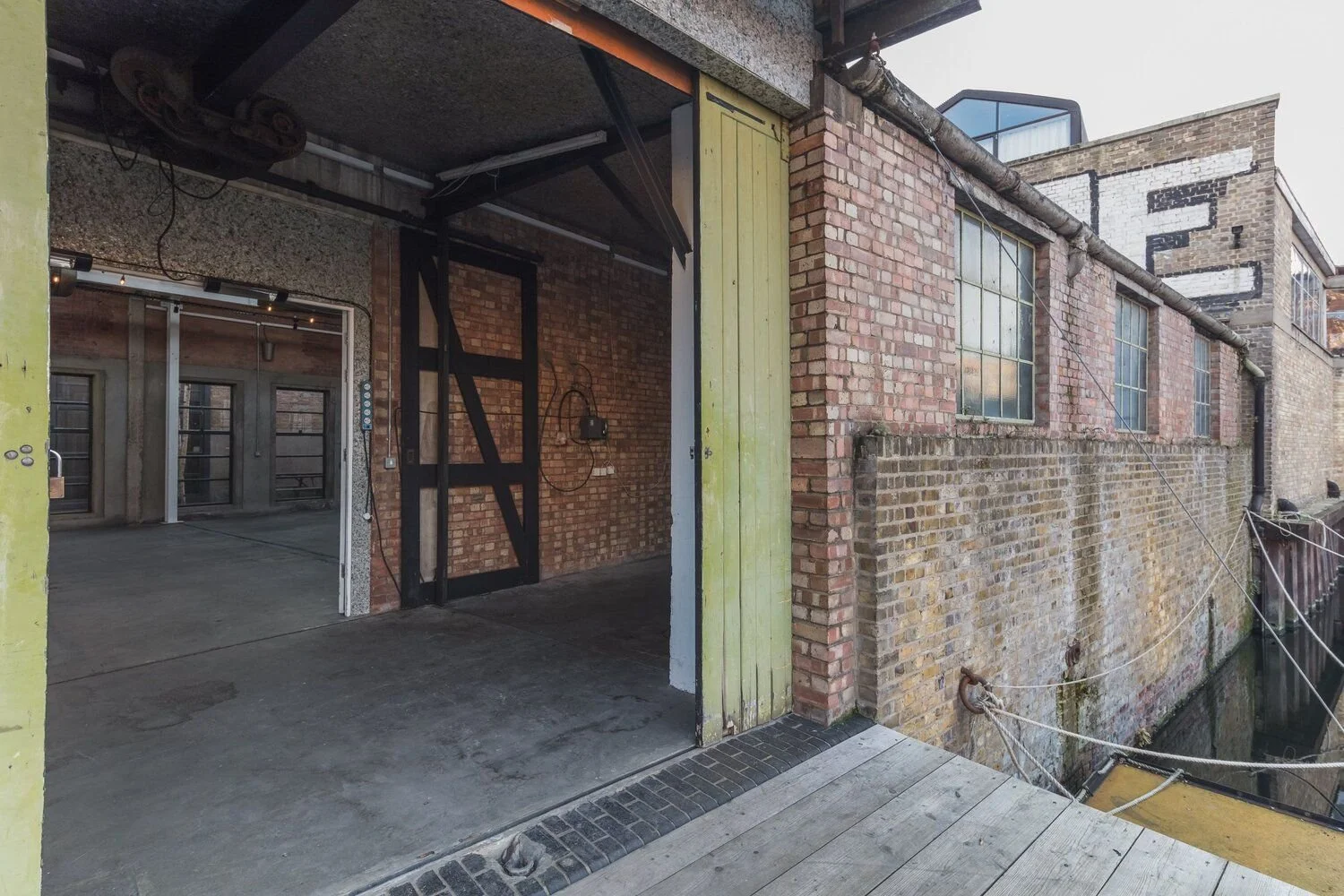 View of an outdoor brick building with windows and a metal drainpipe, featuring an open parking or storage area with a concrete floor and a yellow wooden door, adjacent to a weathered brick wall with a canal below, secured by ropes.