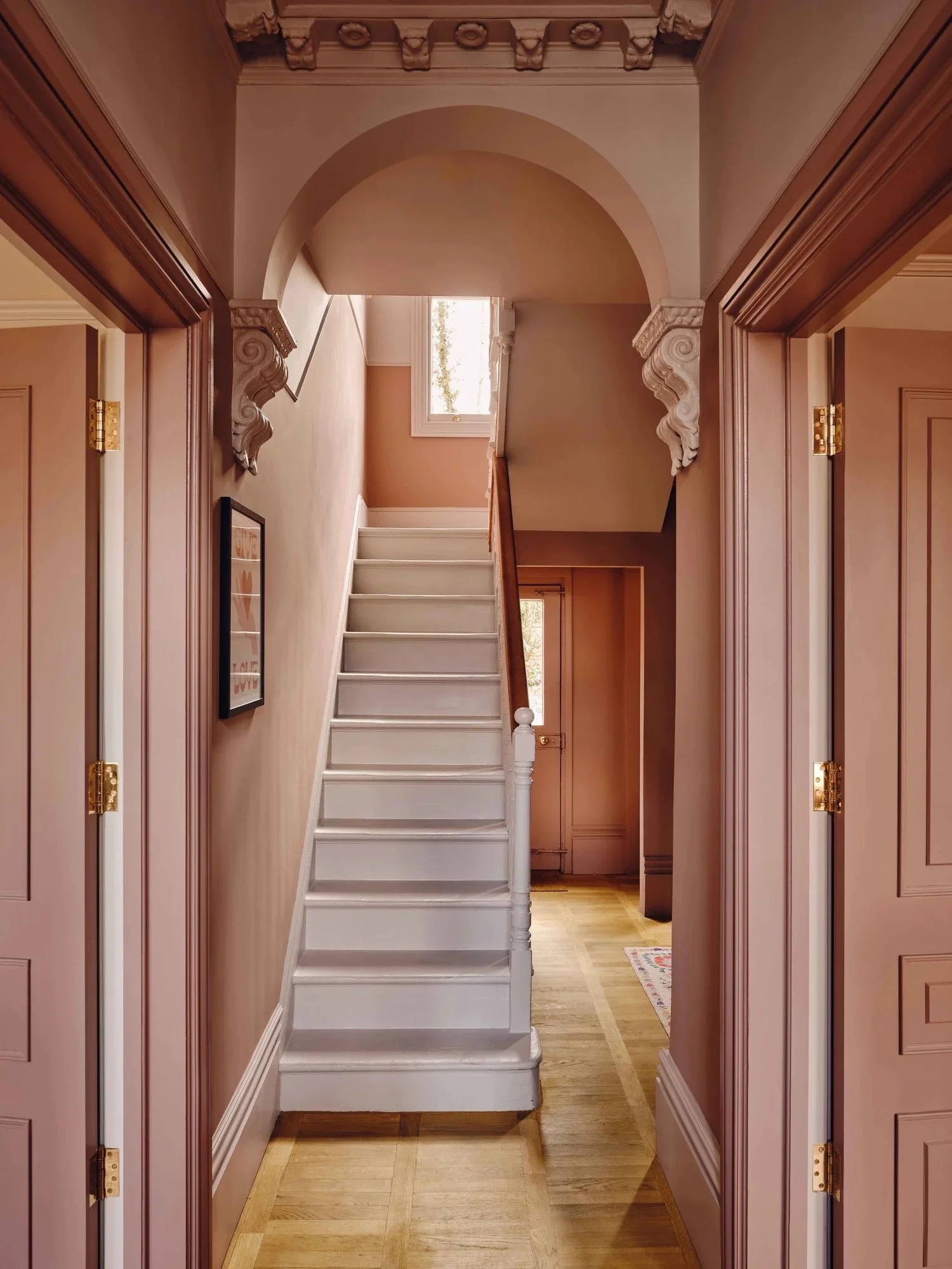 Interior of a house hallway with pink walls, wooden trim, a staircase with white risers and wooden handrail, decorative corbels under the archway, and hardwood floors.