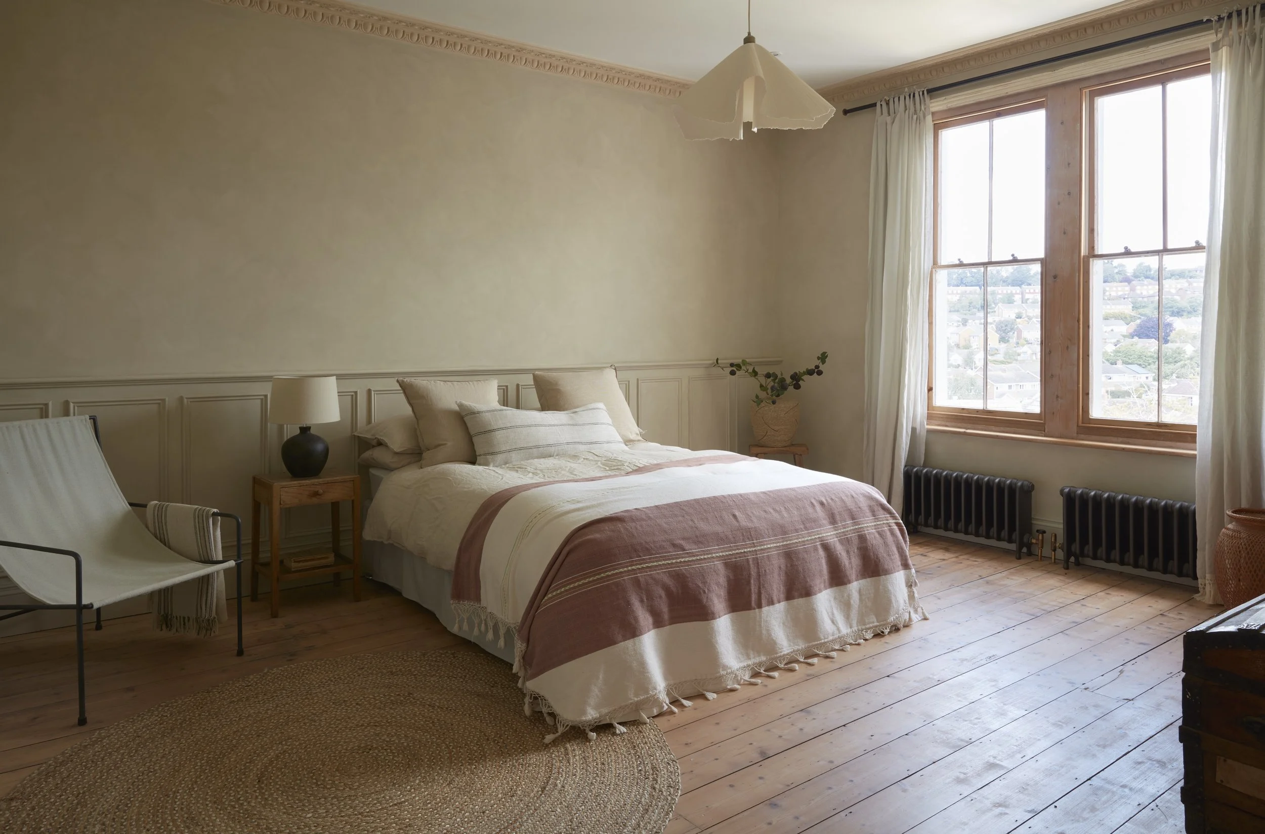 Bright bedroom with a large window, wooden floors, beige walls, a bed with neutral bedding, a small nightstand with a lamp, a white chair, and a potted plant.