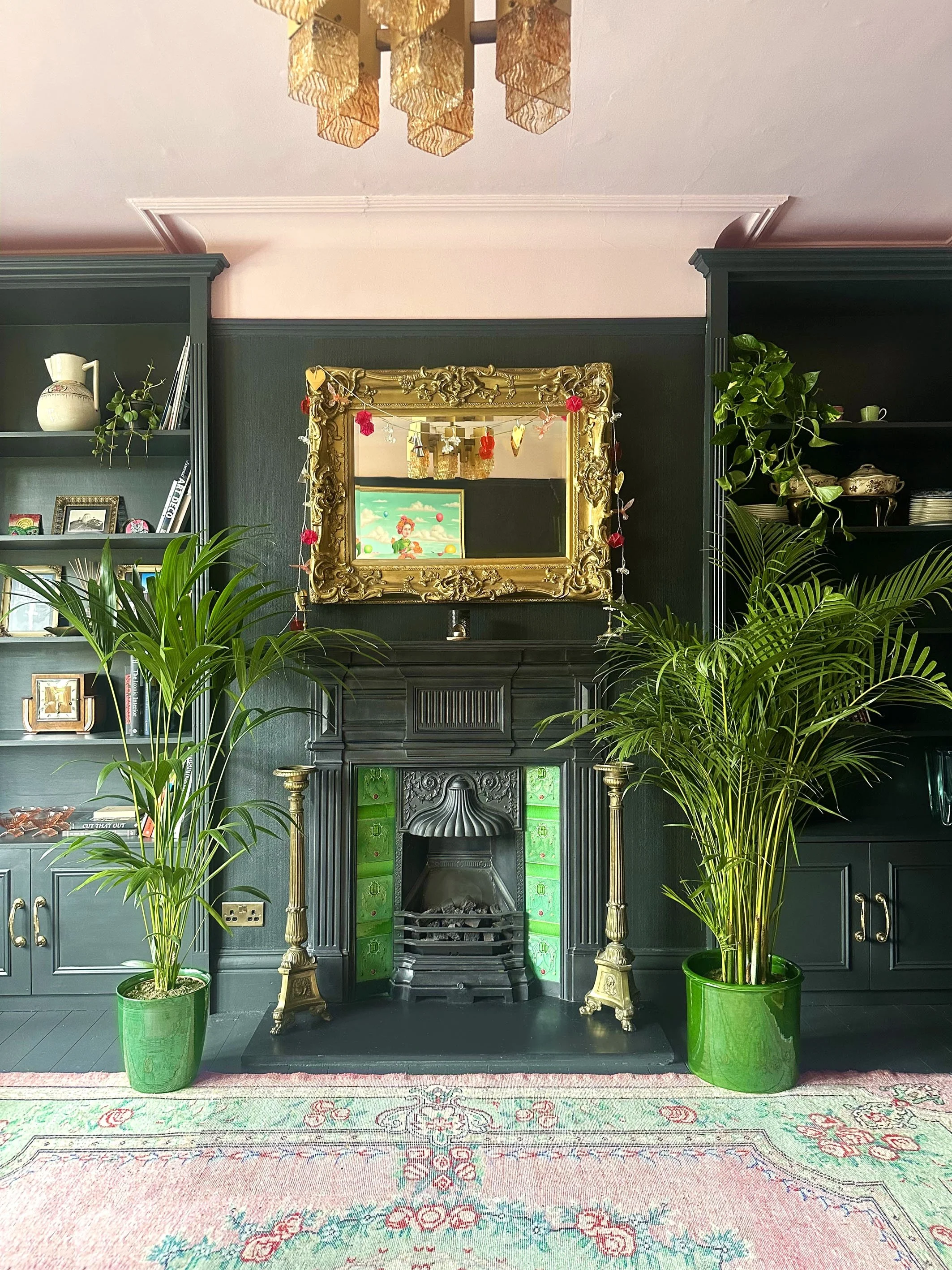 A living room with dark green built-in bookshelves and a black fireplace with colored green and black tiles. The mantel has a large, ornate gold-framed mirror decorated with hanging red and pink paper garlands. Two tall green potted plants are on eit