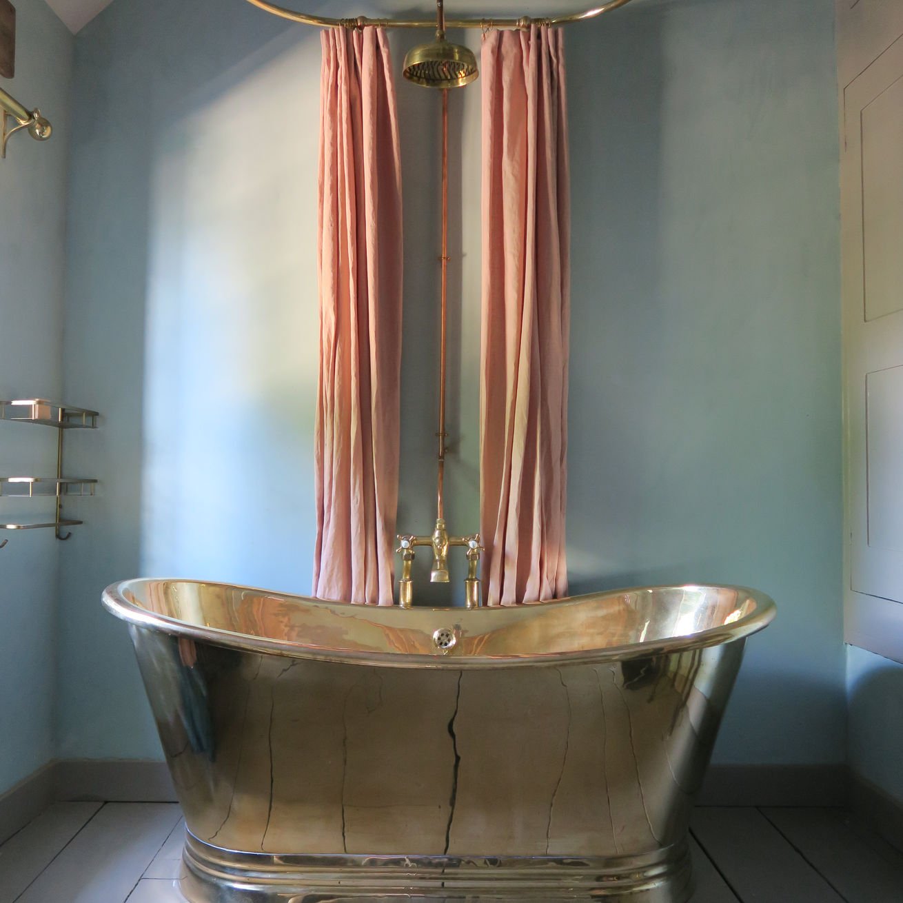 A vintage-style, shiny, metallic bathtub with visible cracks, positioned under a pink curtain, with a blue wall and tiled floor surrounding it.