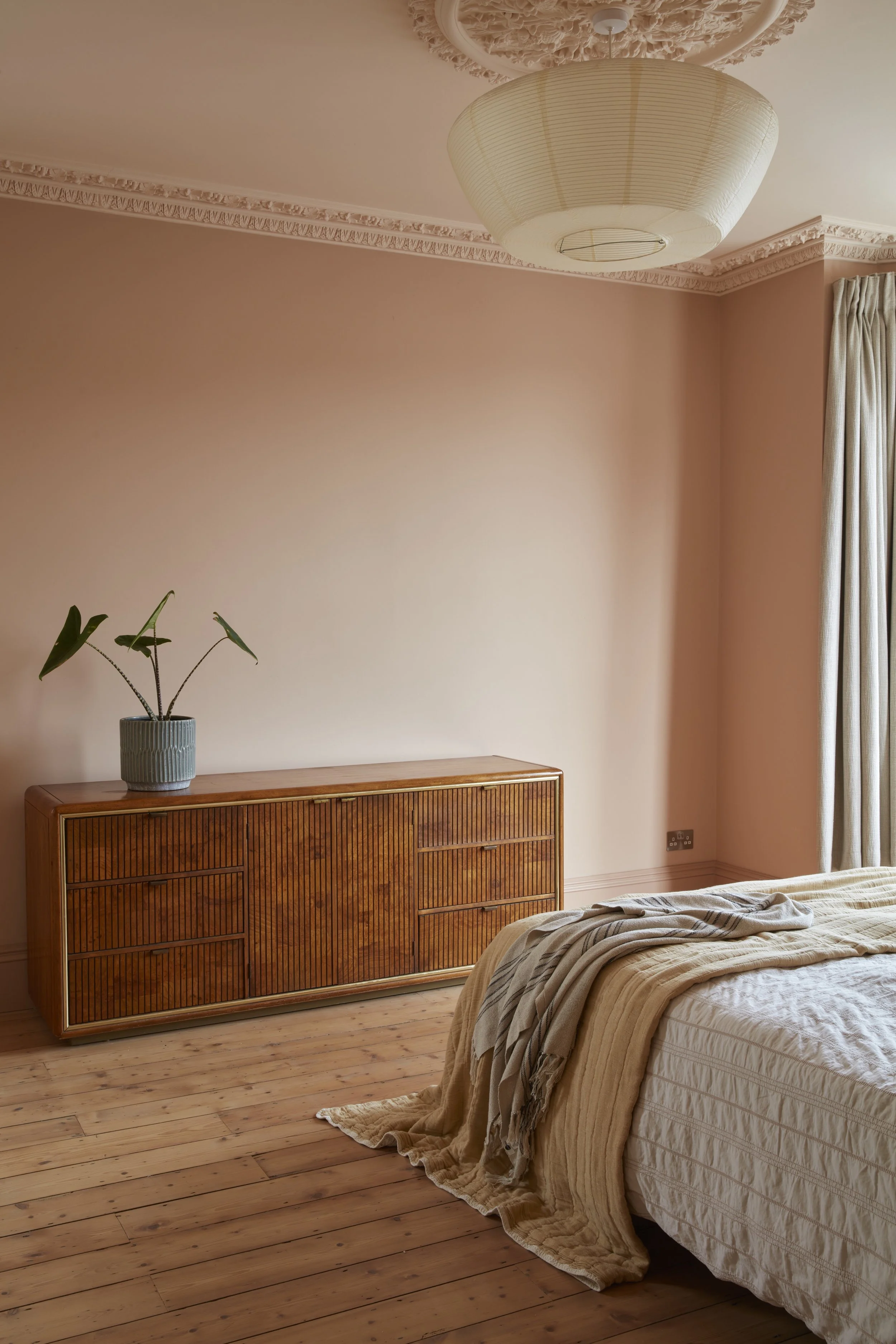A bedroom with wooden flooring, a bed with beige linens, a wooden sideboard, a potted plant, a ceiling light, and a window with curtains.