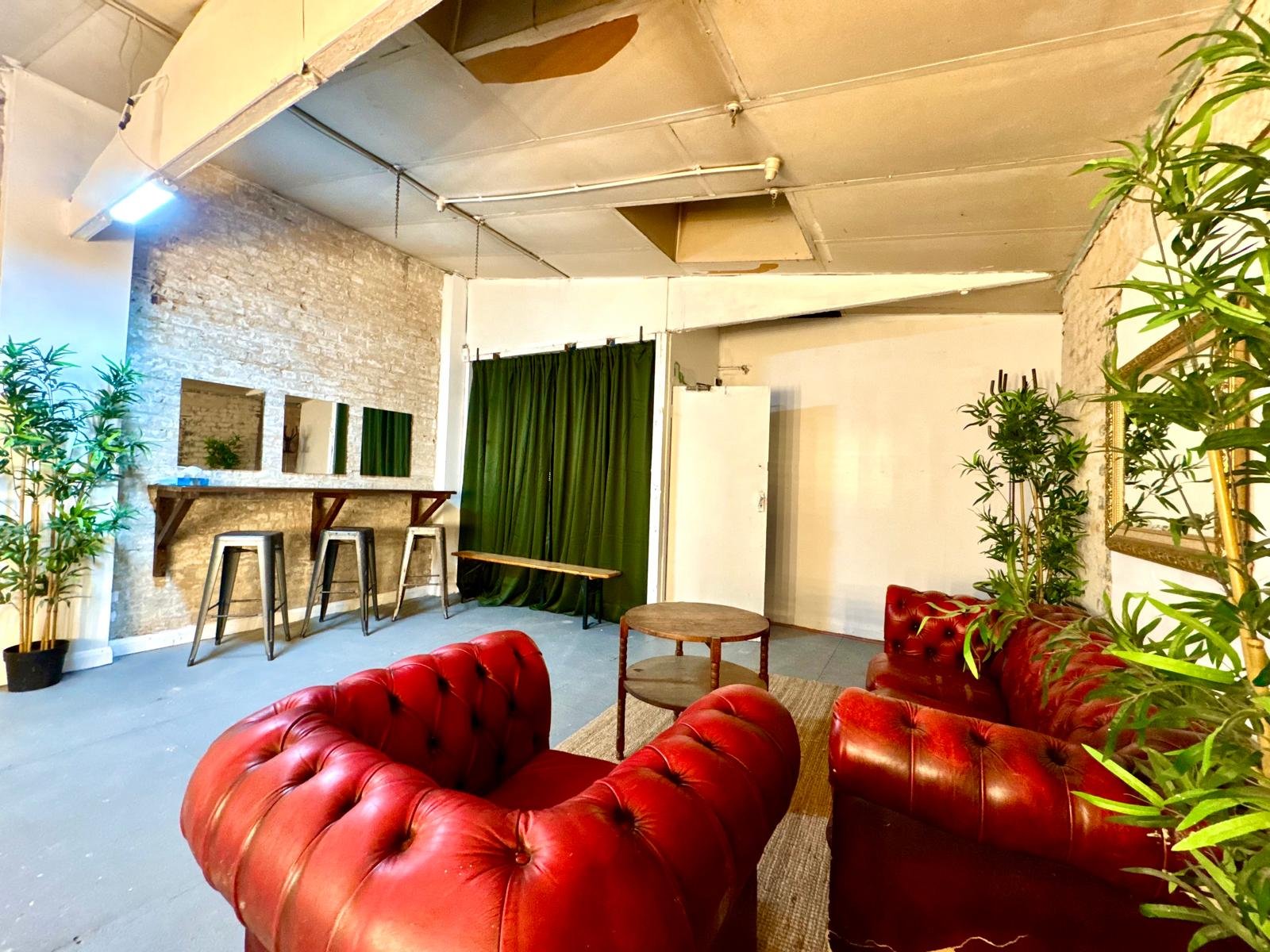 A cozy lounge area with red leather Chesterfield sofas, green plants, a wooden coffee table, a small wooden bench, a white brick wall, a green curtain, and a white door. There are three mirrors on a rustic wooden shelf, and industrial ceiling fixture