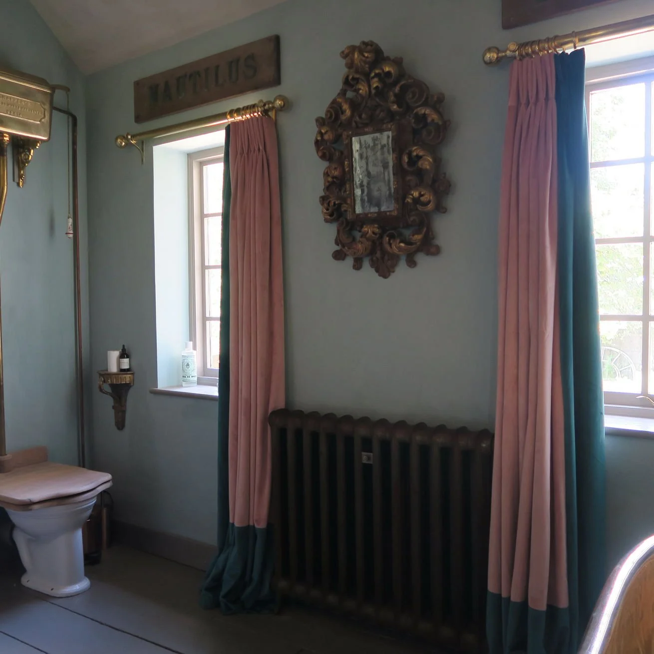 Interior of a vintage-style room with two small windows, pink and teal curtains, a wooden mirror with ornate carvings, a wooden sign above one window, and a radiator underneath the mirror.