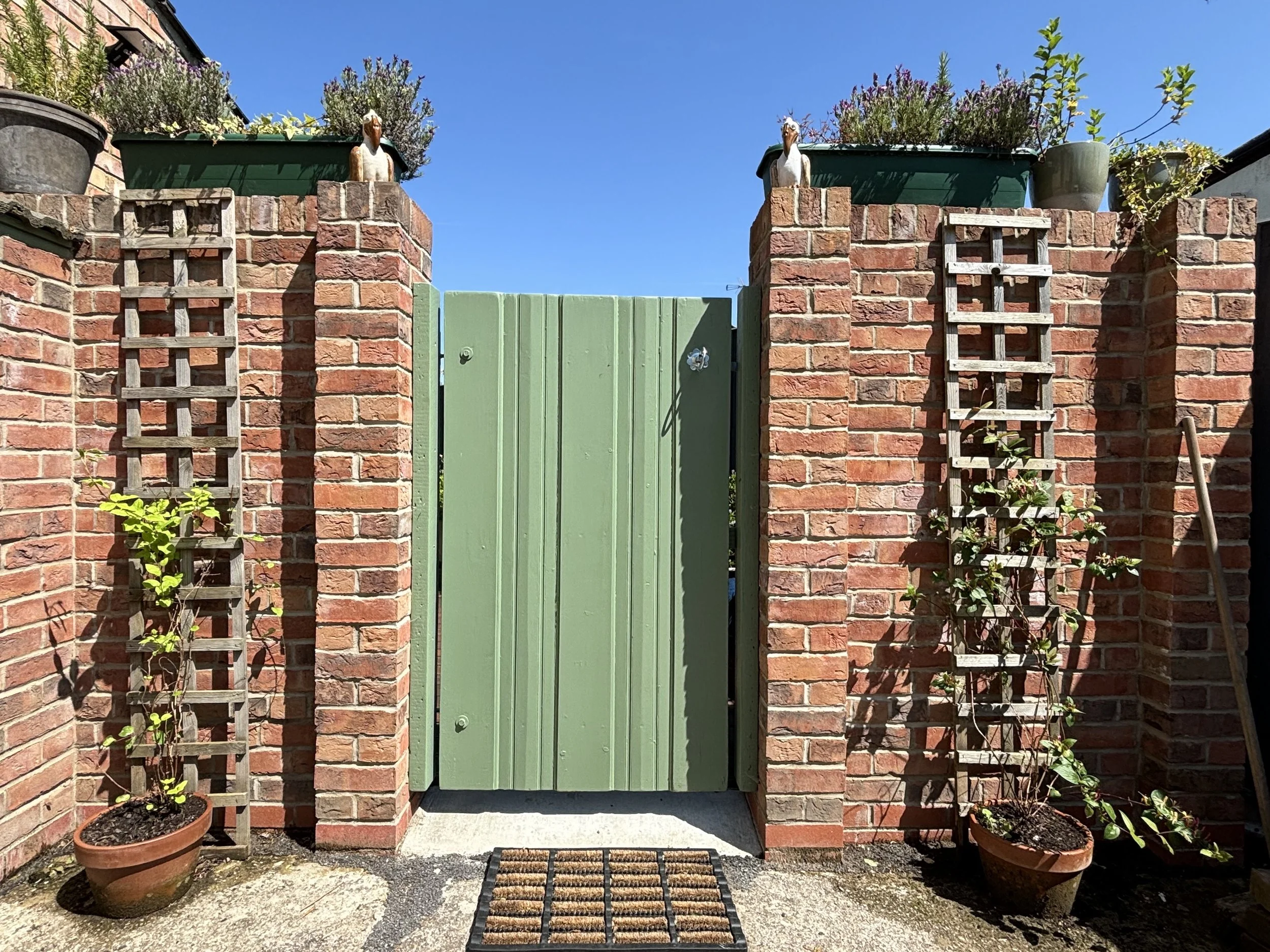 Green wooden gate set between brick walls with climbing plants, with potted plants on either side and decorative garden statues on top of the walls.