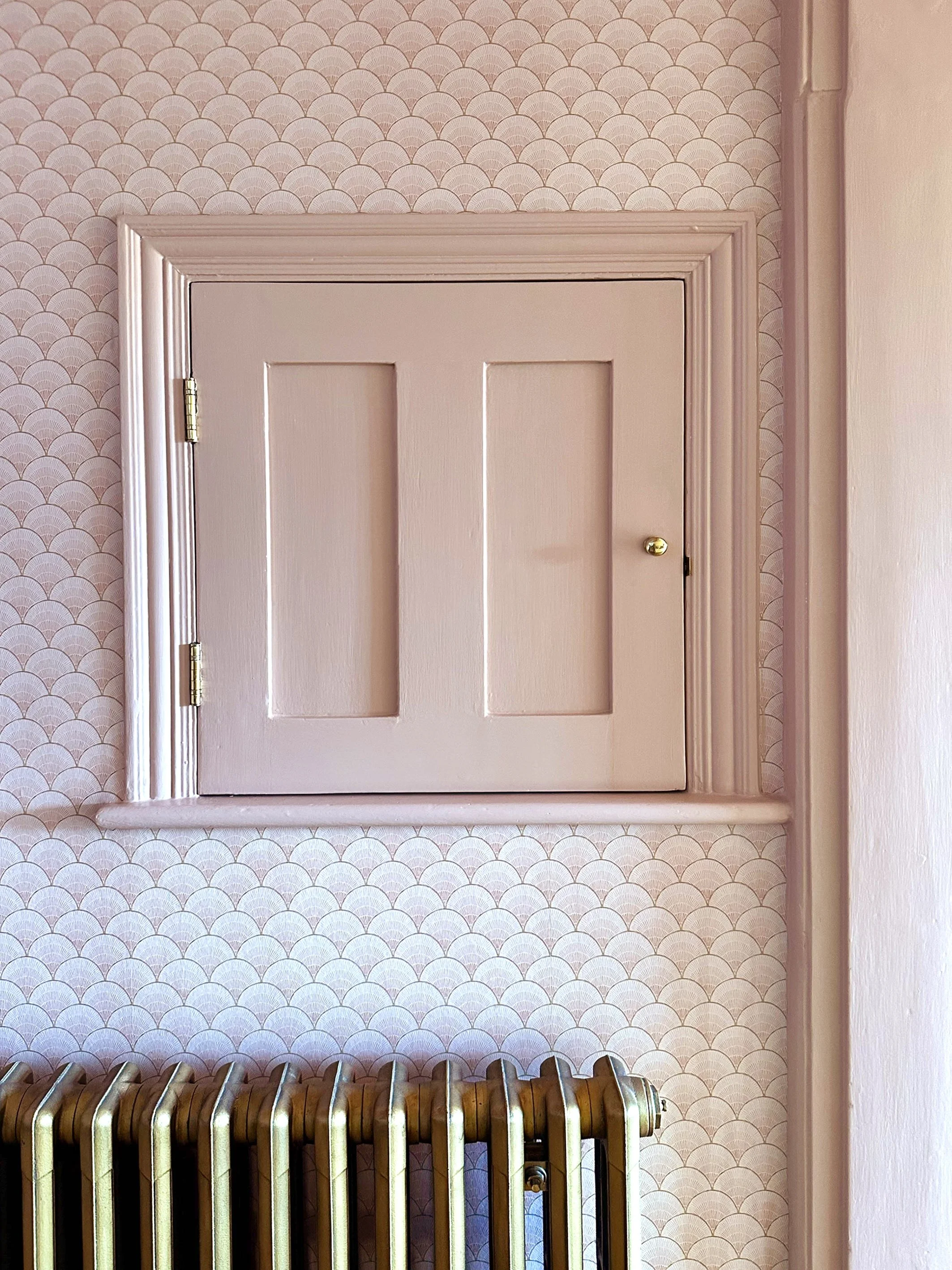 A small pink cabinet with a round doorknob, mounted on a wall with pink scallop-patterned wallpaper and a brass radiator below.