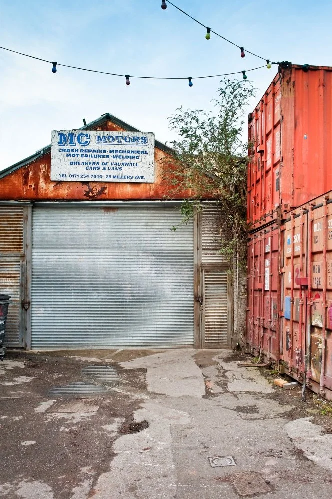 Small auto repair shop with a faded blue and white sign that reads "MC MOTORS, Auto Repairs, Mechanical, MOT Failures, Welding, Breakers of Vauxhall, Cars & Vans, Tel 0171 254 7640, 28 Millers Ave." with a rusty metal roller shutter door, surrounded 