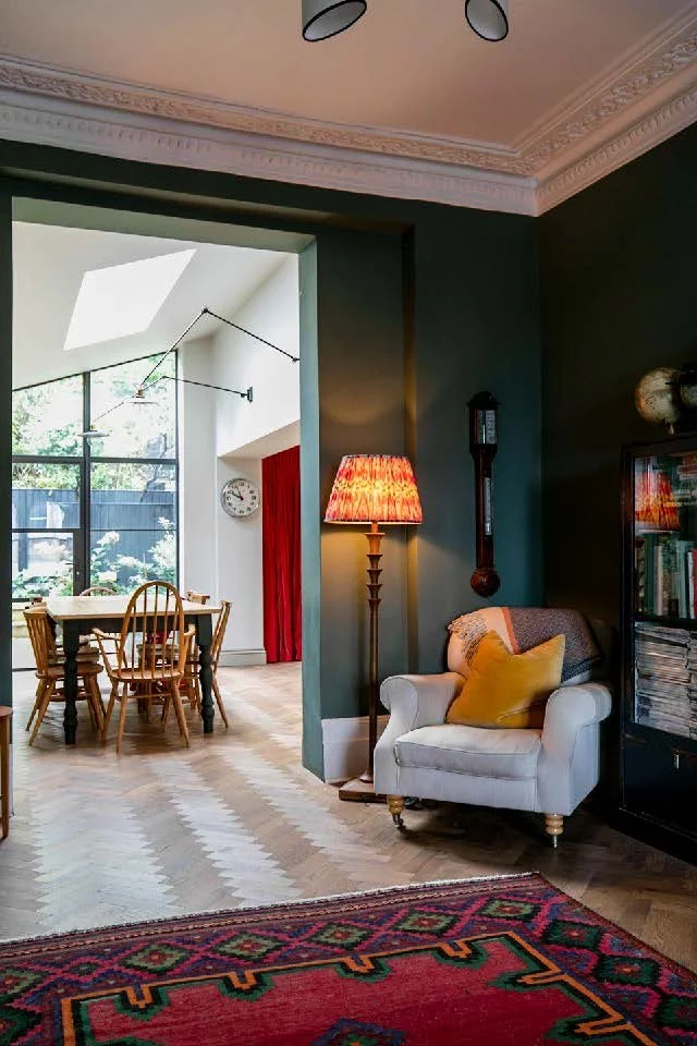 Living room and dining area with large windows, hardwood floors, a white armchair with a yellow pillow, a tall lamp with a colorful shade, and a bookshelf.