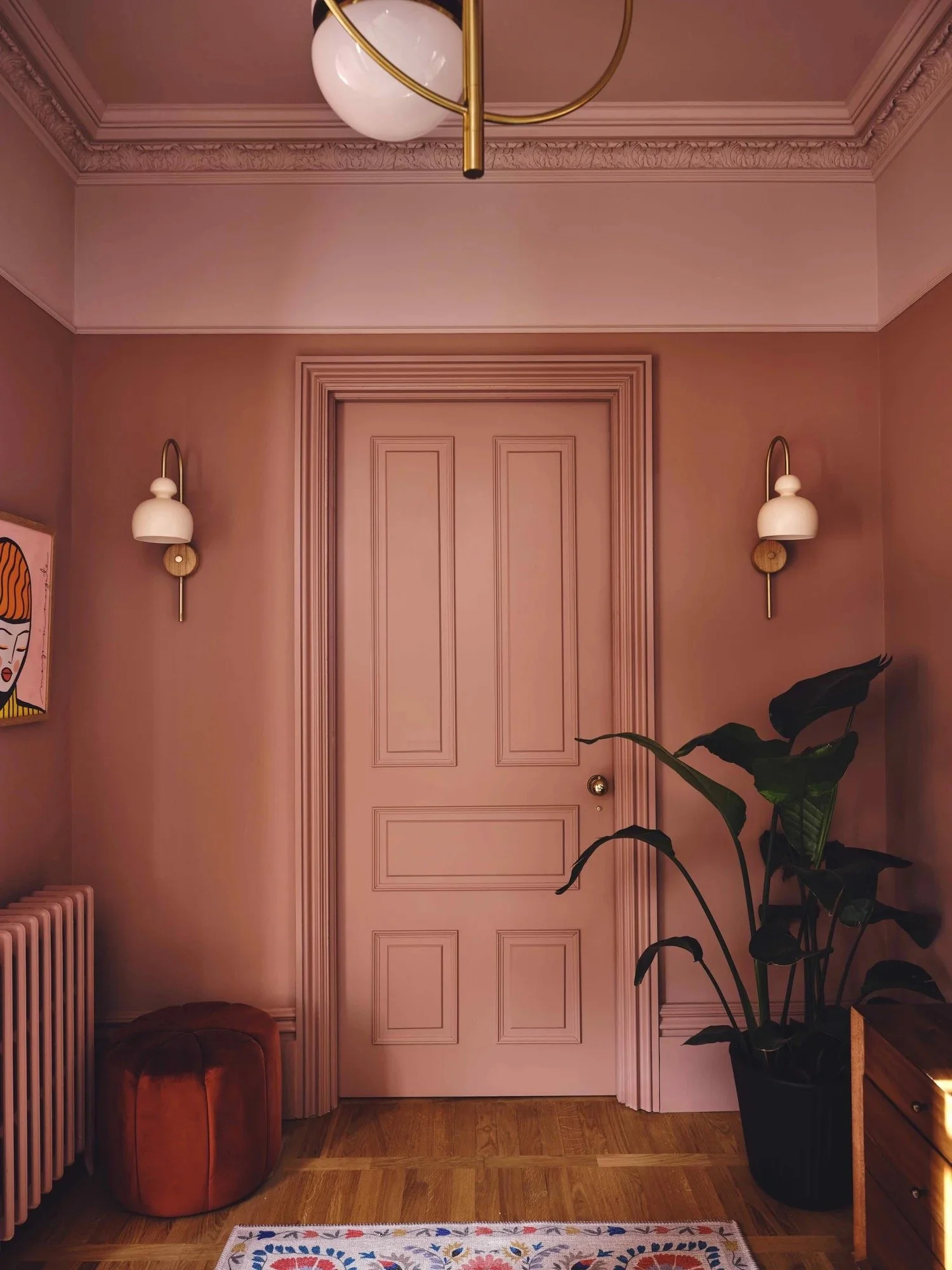 Interior view of a room with pink walls and a pink door, decorative crown molding, and two wall-mounted lamps. There is a large potted plant on the right, a round orange ottoman on the left, a colorful rug on the floor, and part of a colorful art pie