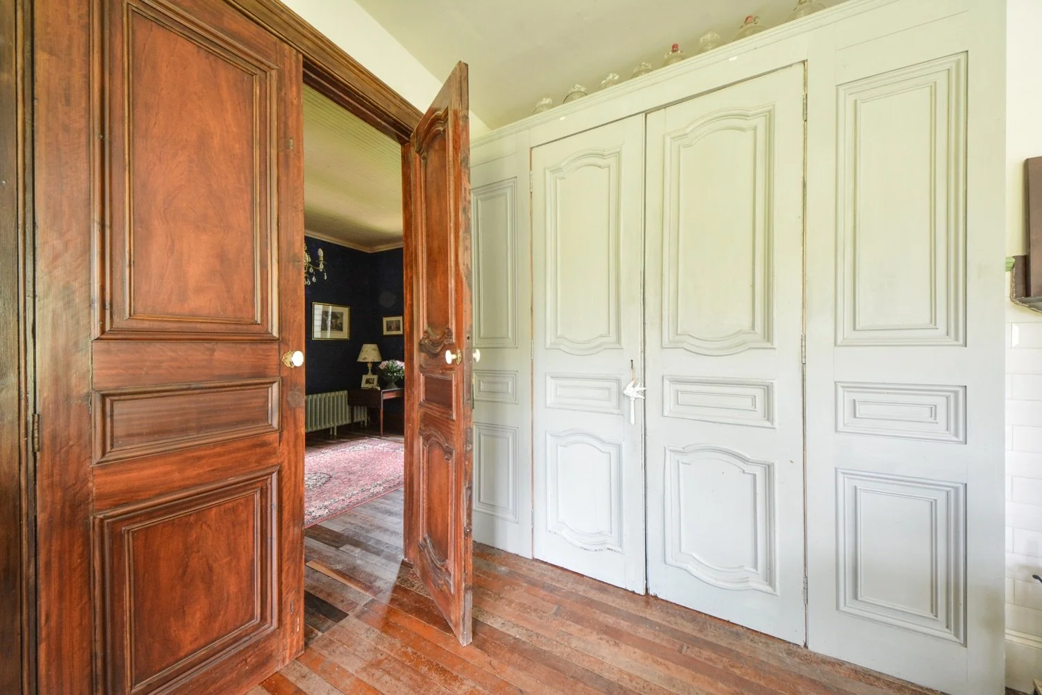 Interior view of a room with a wooden double door open to a living space with dark walls, a chandelier, framed pictures, a lamp, and a rug. There is a white closet with paneled doors on the right side.