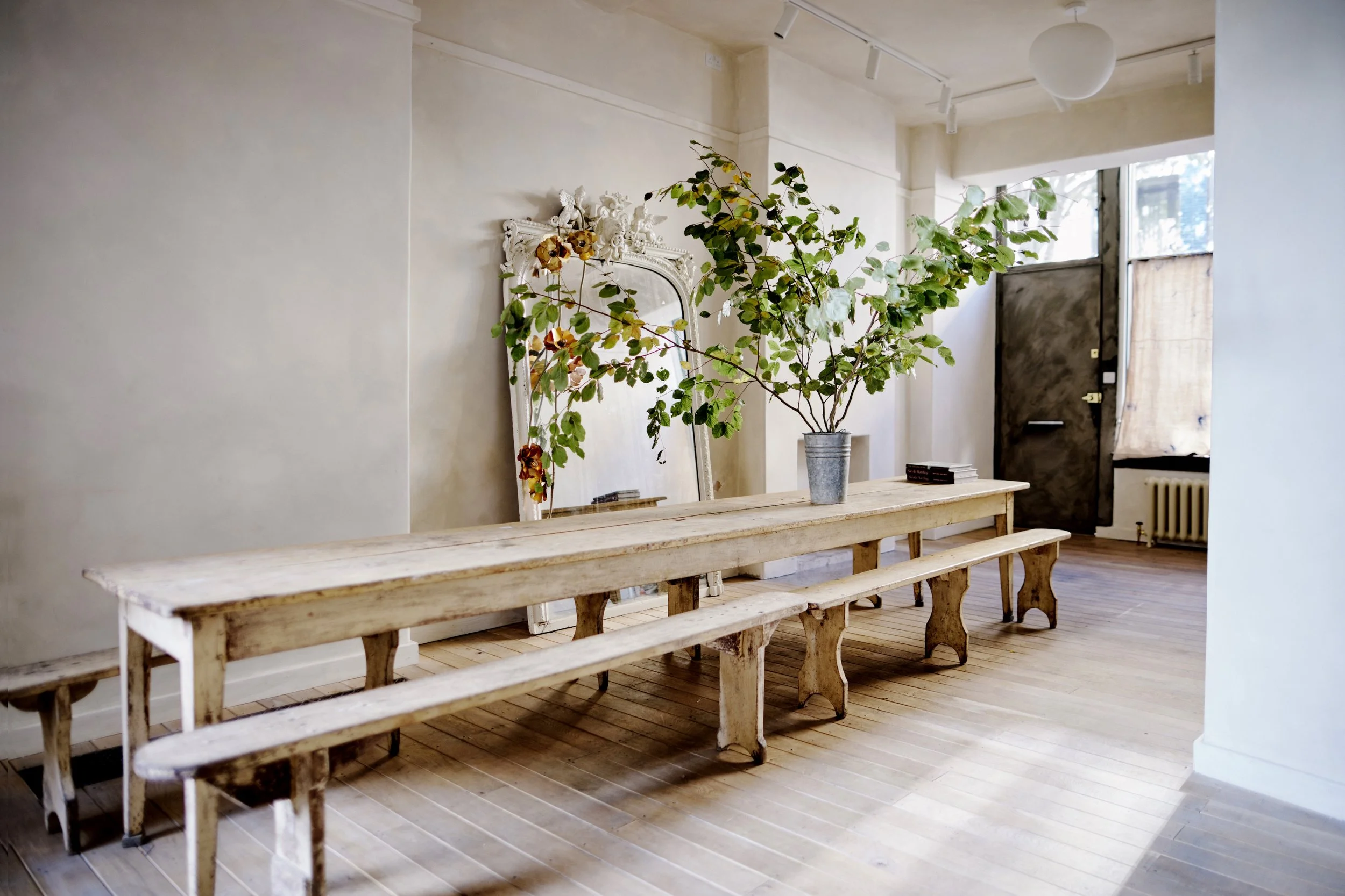 Empty rustic wooden table with matching benches next to a large potted plant in a room with hardwood floors, a decorative mirror, and an industrial-style door near a window.