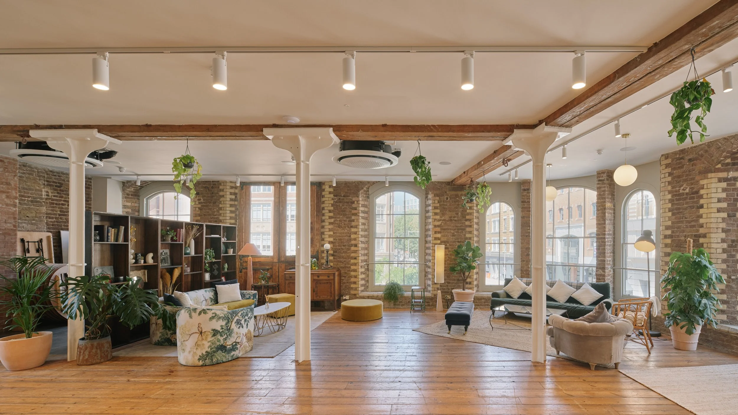 Spacious living room with exposed brick walls, large arched windows, wooden beams, and a mix of vintage and modern furniture, decorated with potted plants and shelving.