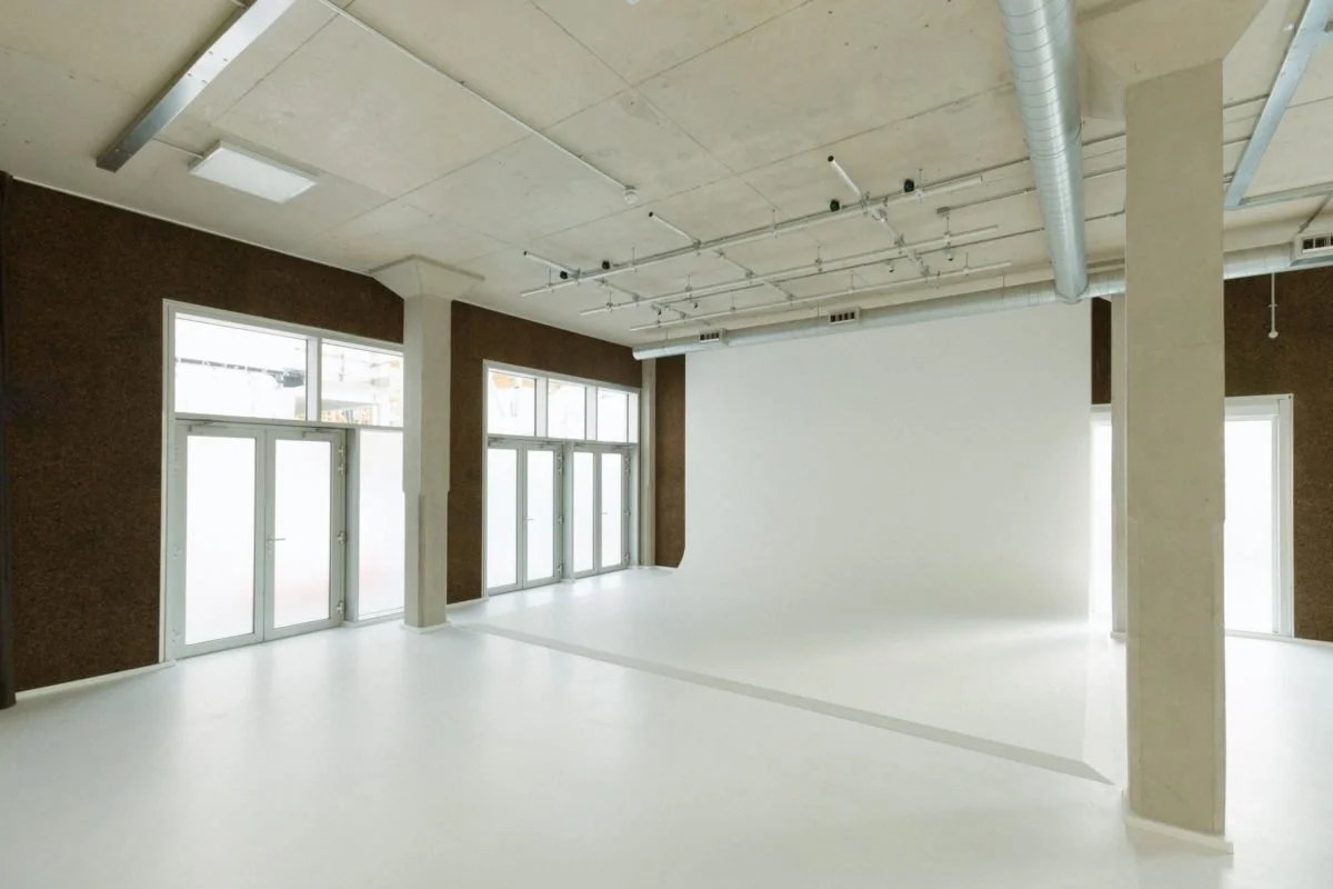 Empty room with large windows, white flooring, and exposed ceiling with pipes and lighting fixtures.