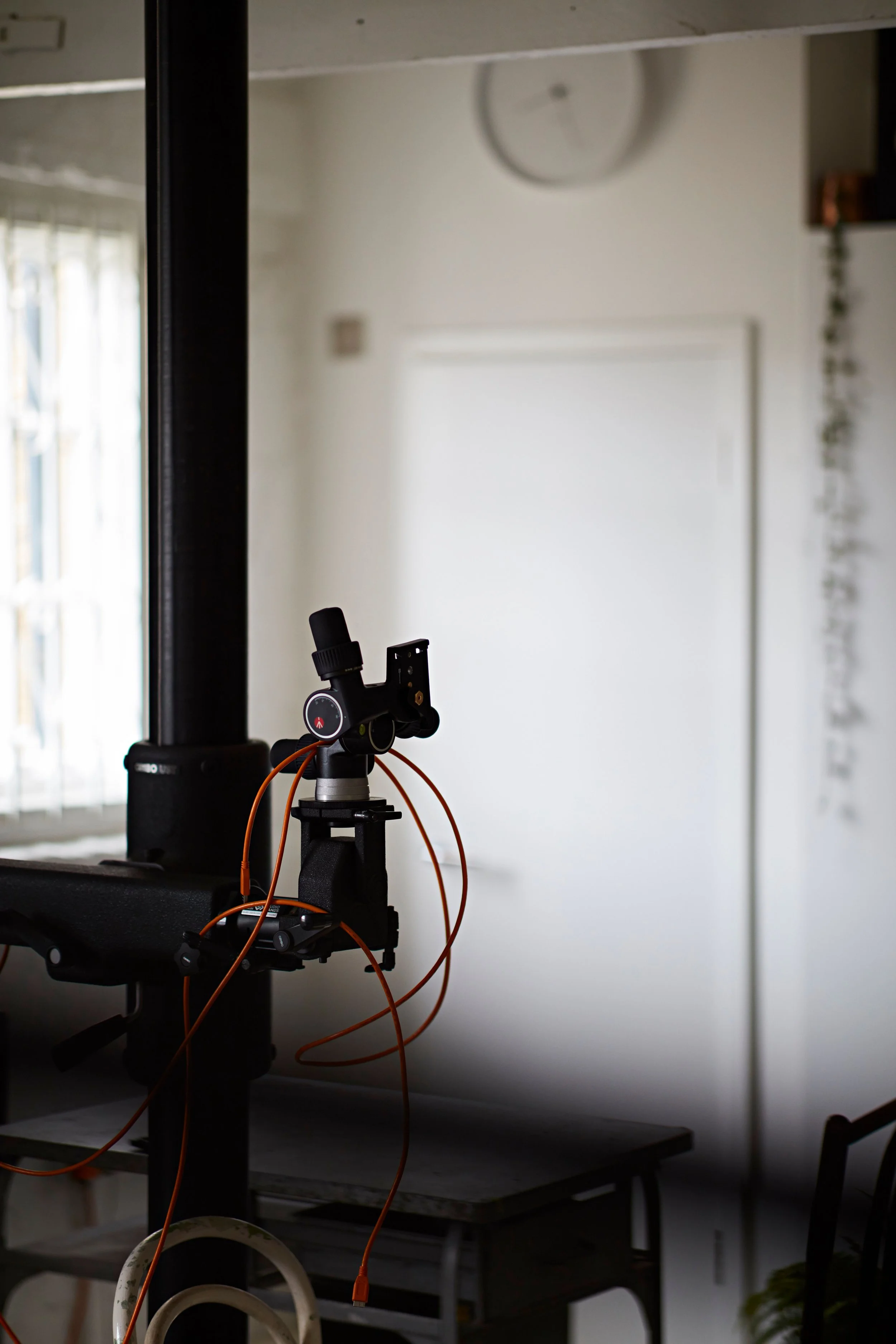 A camera mounted on a tripod in a room, with orange cables hanging from it. The room has white walls, a white door or panel, a wall clock, and some furniture and decor.