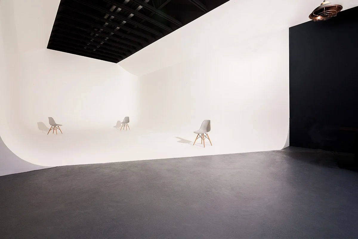 Empty photography studio with three white chairs arranged on a seamless white backdrop, black ceiling, and dark floor.