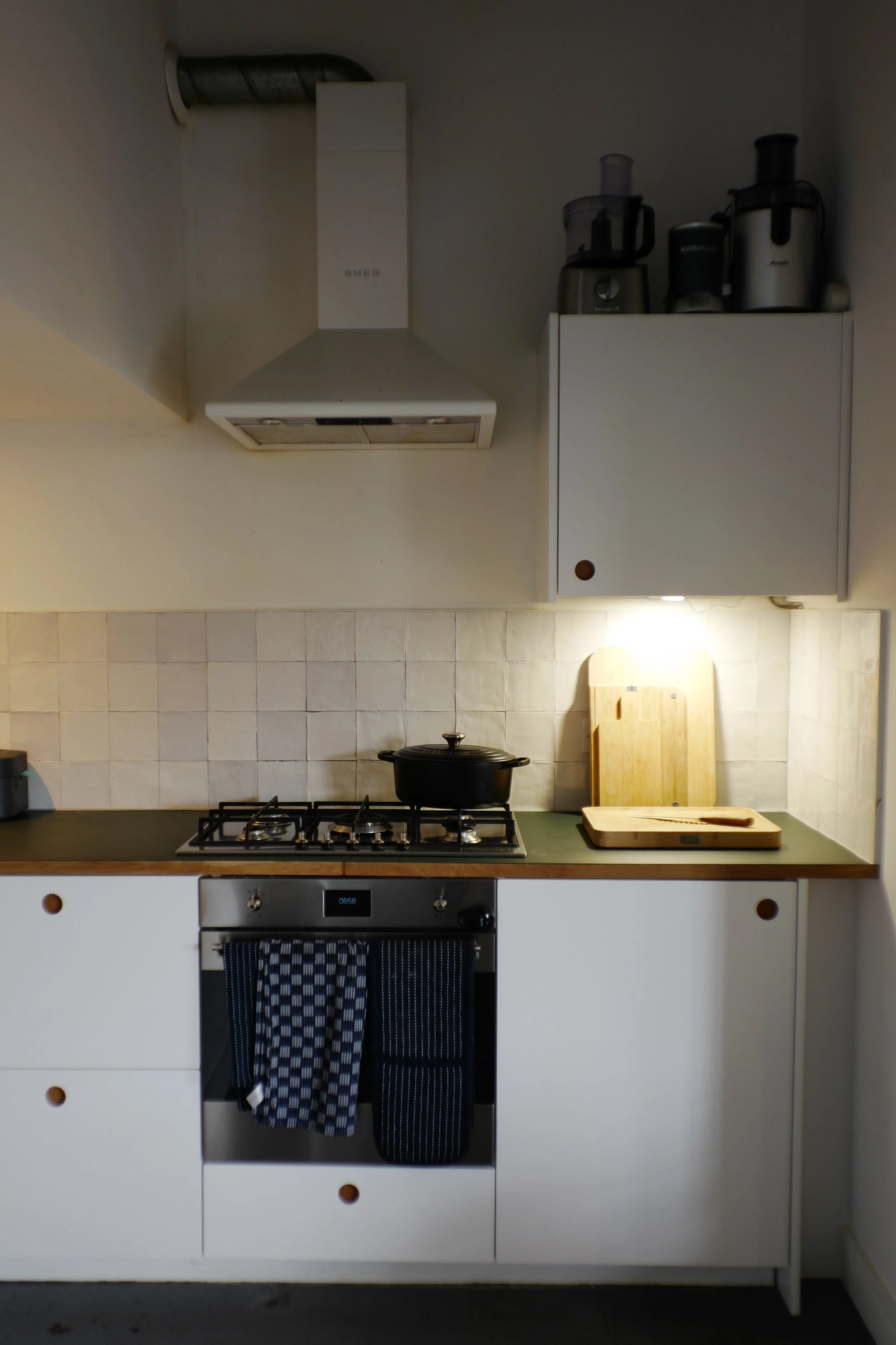 A kitchen with white cabinets, a stove with a black pot, a wooden countertop with cutting boards, a range hood, and kitchen appliances on a white wall cabinet.