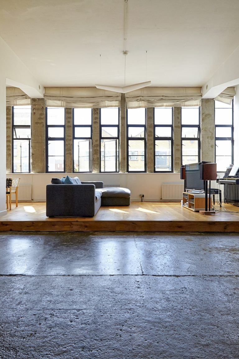 Empty loft with large windows, exposed concrete, a gray sofa, and a TV setup on a wooden platform.