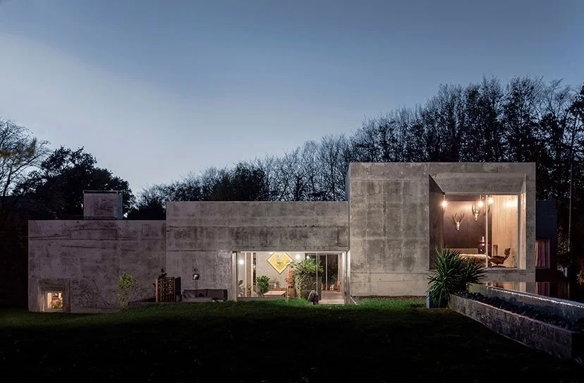 Modern concrete house during twilight with visible indoor lighting and large glass doors, surrounded by a grassy yard and trees in the background.