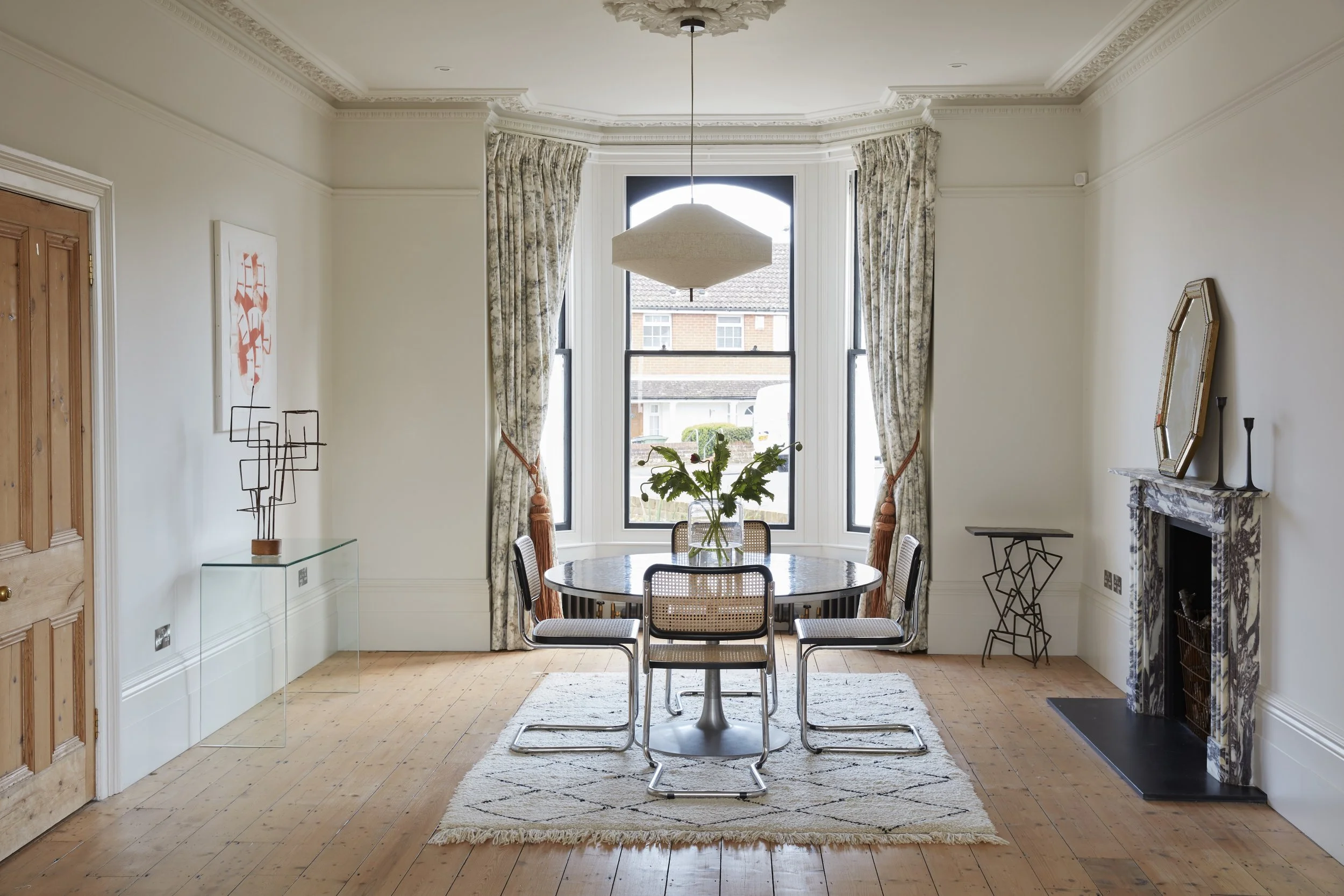 A dining room with a round table, six chairs, a large window with floral curtains, a decorative rug, a fireplace with a mirror, and artwork on the white walls.
