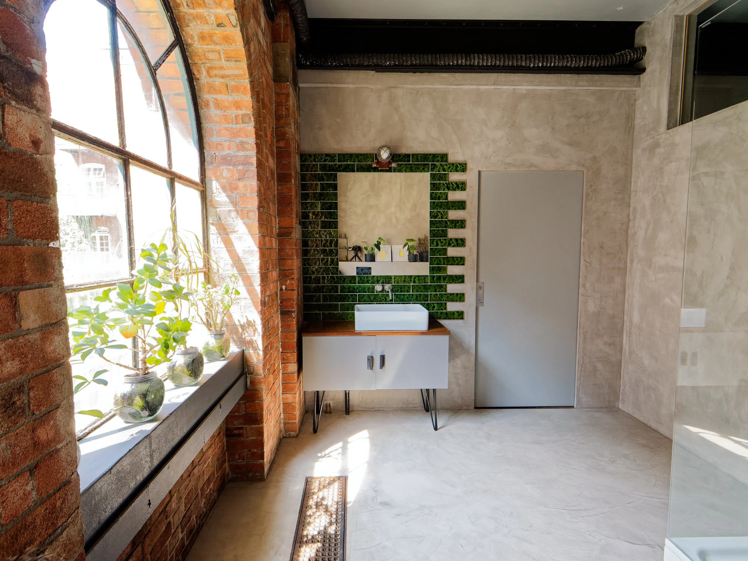 Modern bathroom with exposed brick wall, large window with potted plants, and a green tiled wall with a mirror and sink.