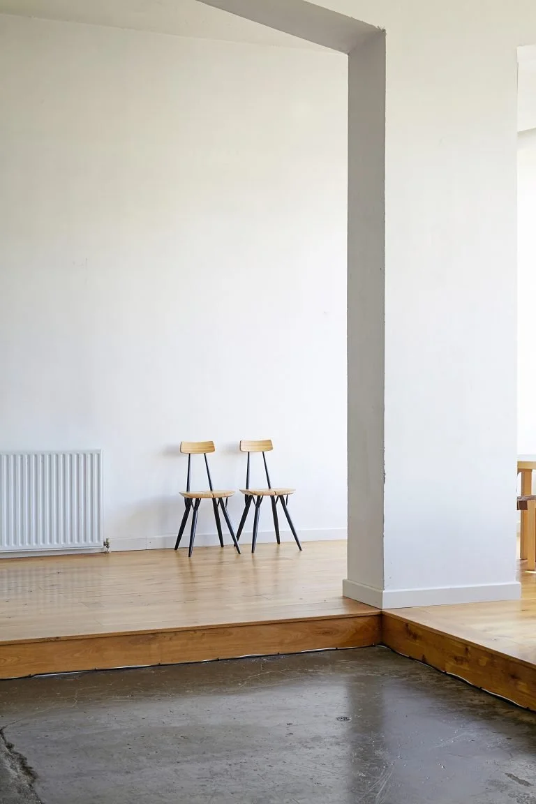 Two wooden chairs with black legs positioned against a white wall in a minimalistic room, with a radiator on the left side and a small step leading to a different floor level.