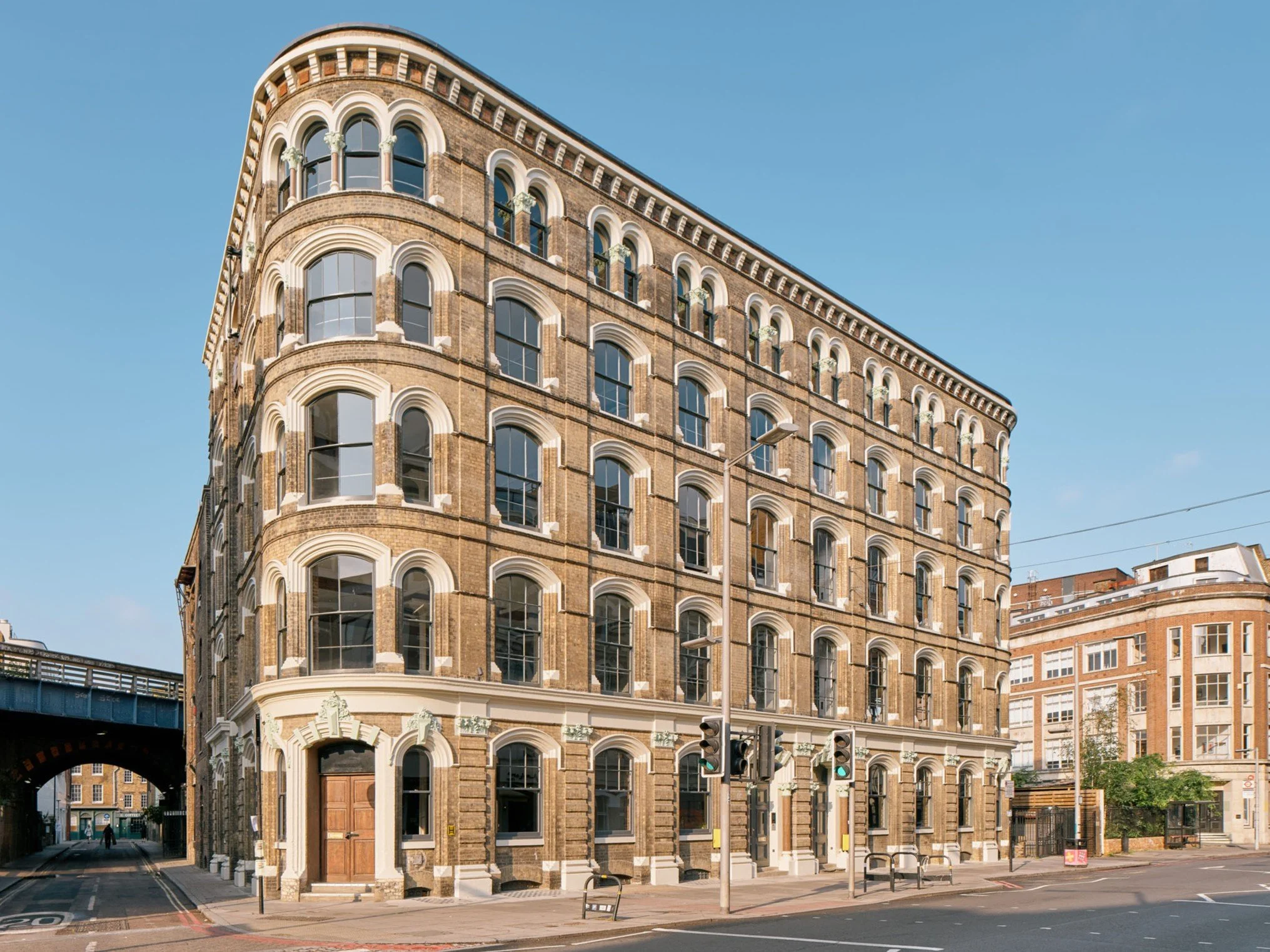 A historic brick building with arched windows and decorative white trim on a city street corner, with traffic lights and a clear blue sky.