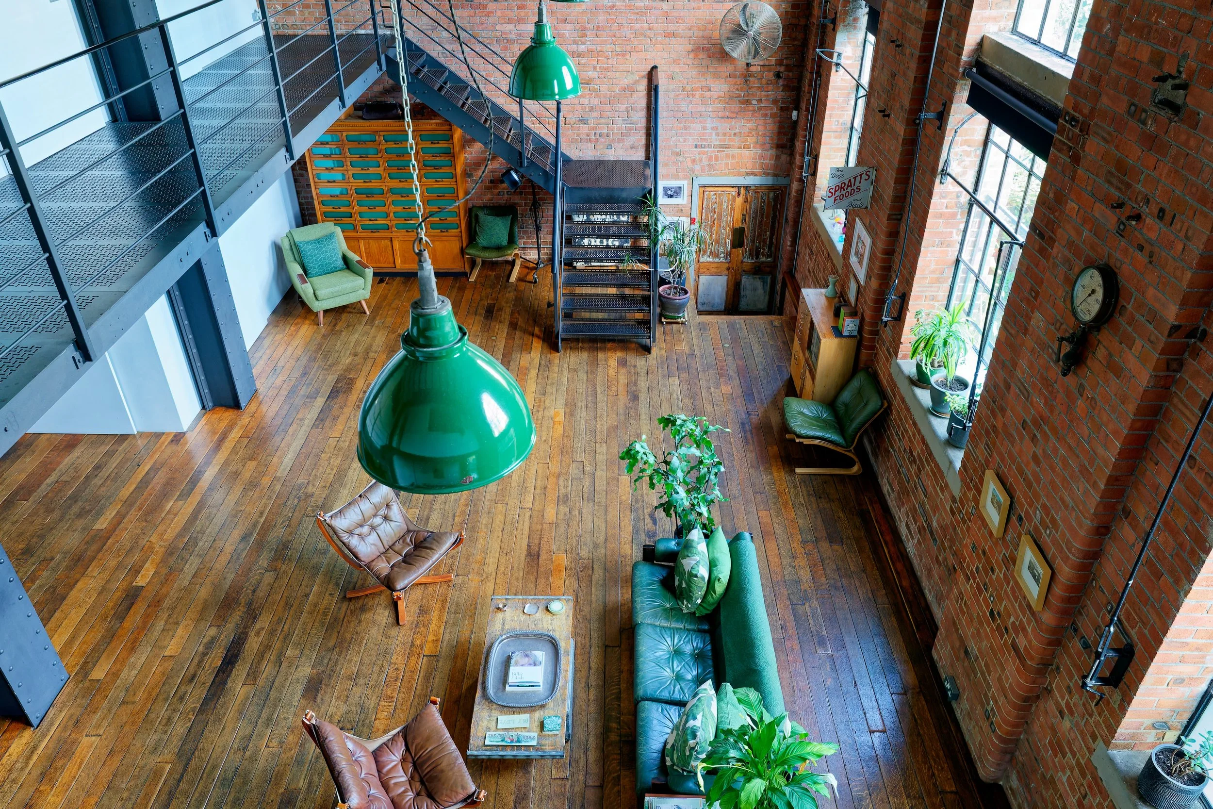 Interior of a loft-style living space with wooden flooring, brick walls, large windows with potted plants, green pendant lights, leather chairs, a green sofa, and a staircase leading to a mezzanine.