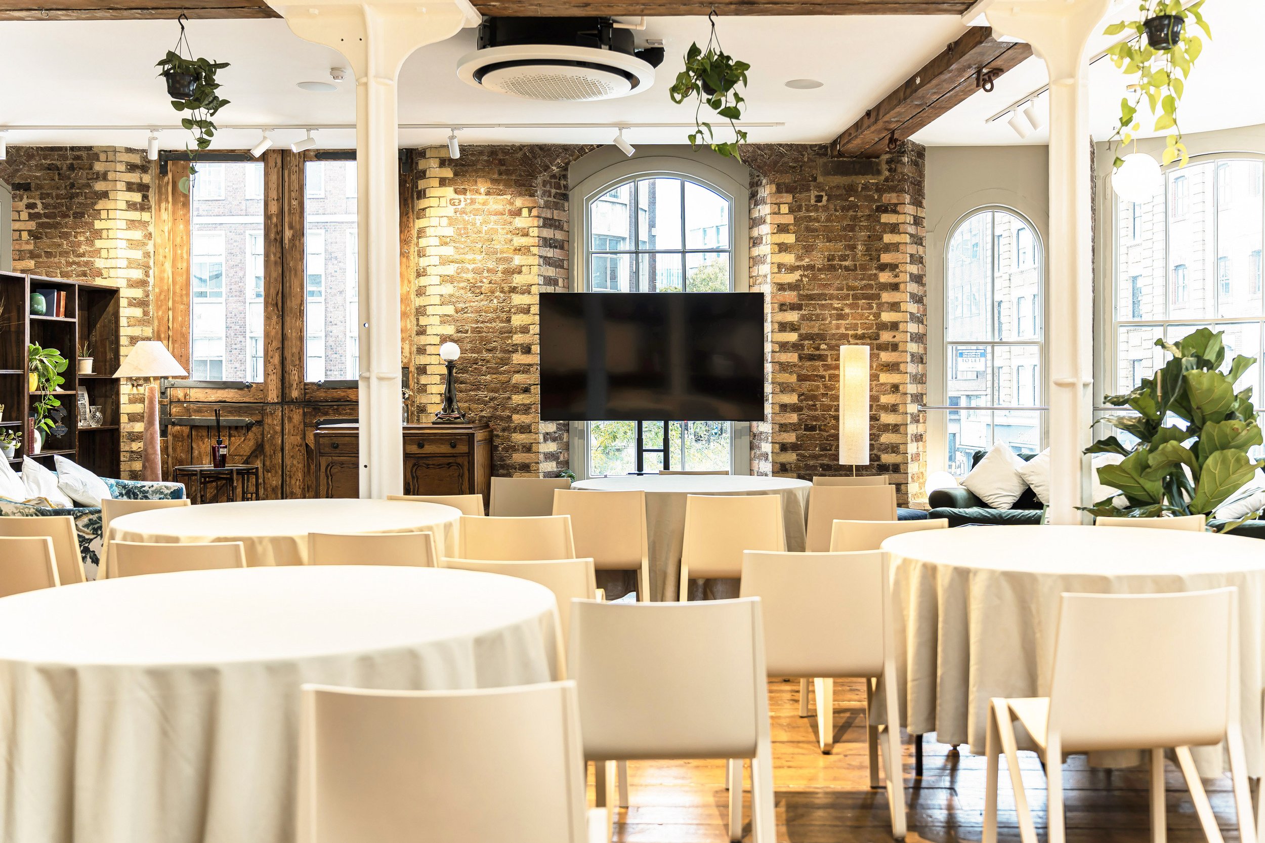 A modern, cozy event space or conference room with round tables covered in beige tablecloths, surrounded by white chairs. The room features exposed brick walls, large arched windows allowing natural light, and decorative greenery hanging from the cei