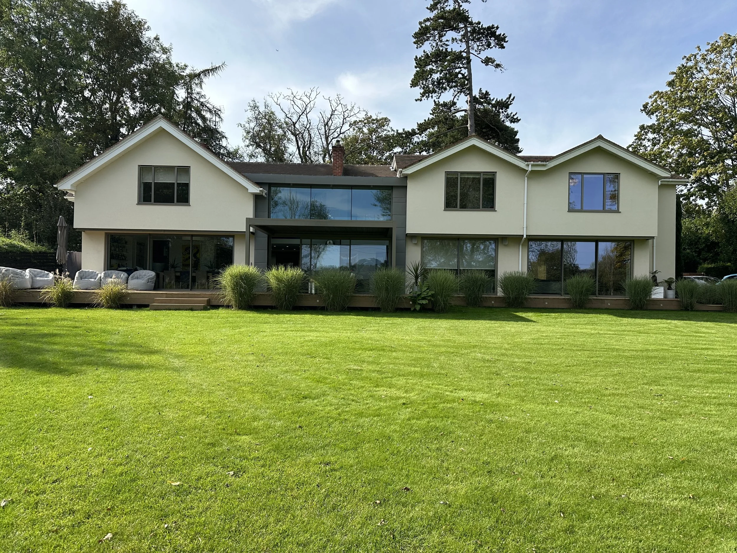 Modern two-story house with large windows, a wooden deck with outdoor furniture, and a green lawn in front.