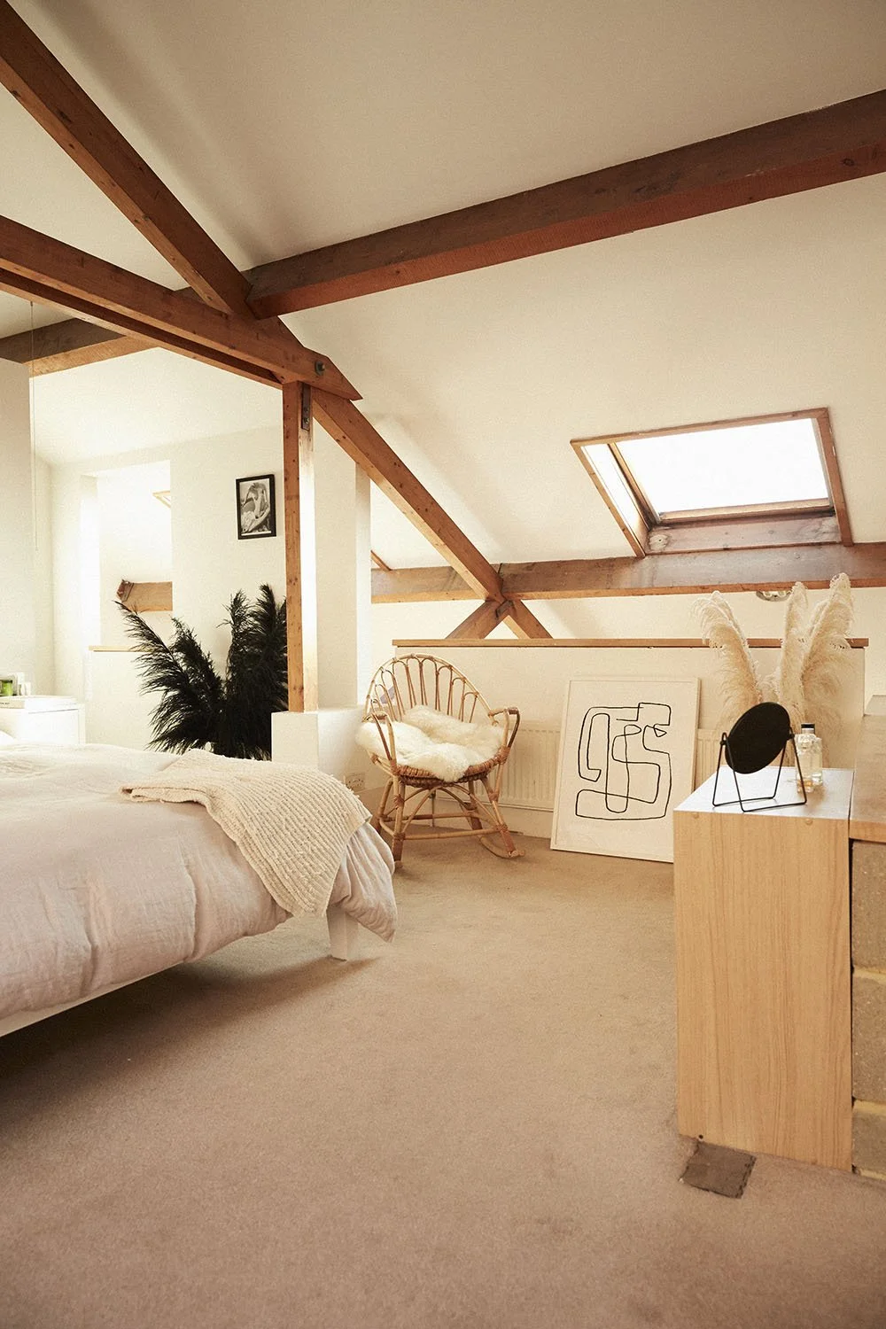 A cozy attic bedroom with a bed, a wicker chair with a pillow, framed art, decorative plants, and natural light coming through skylight windows.