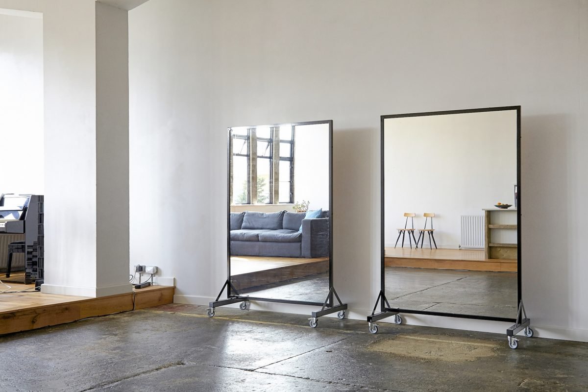 Two full-length mirrors on wheels in front of a white wall, with a living room with a gray couch reflected in one mirror and a dining area with two chairs and a counter reflected in the other.