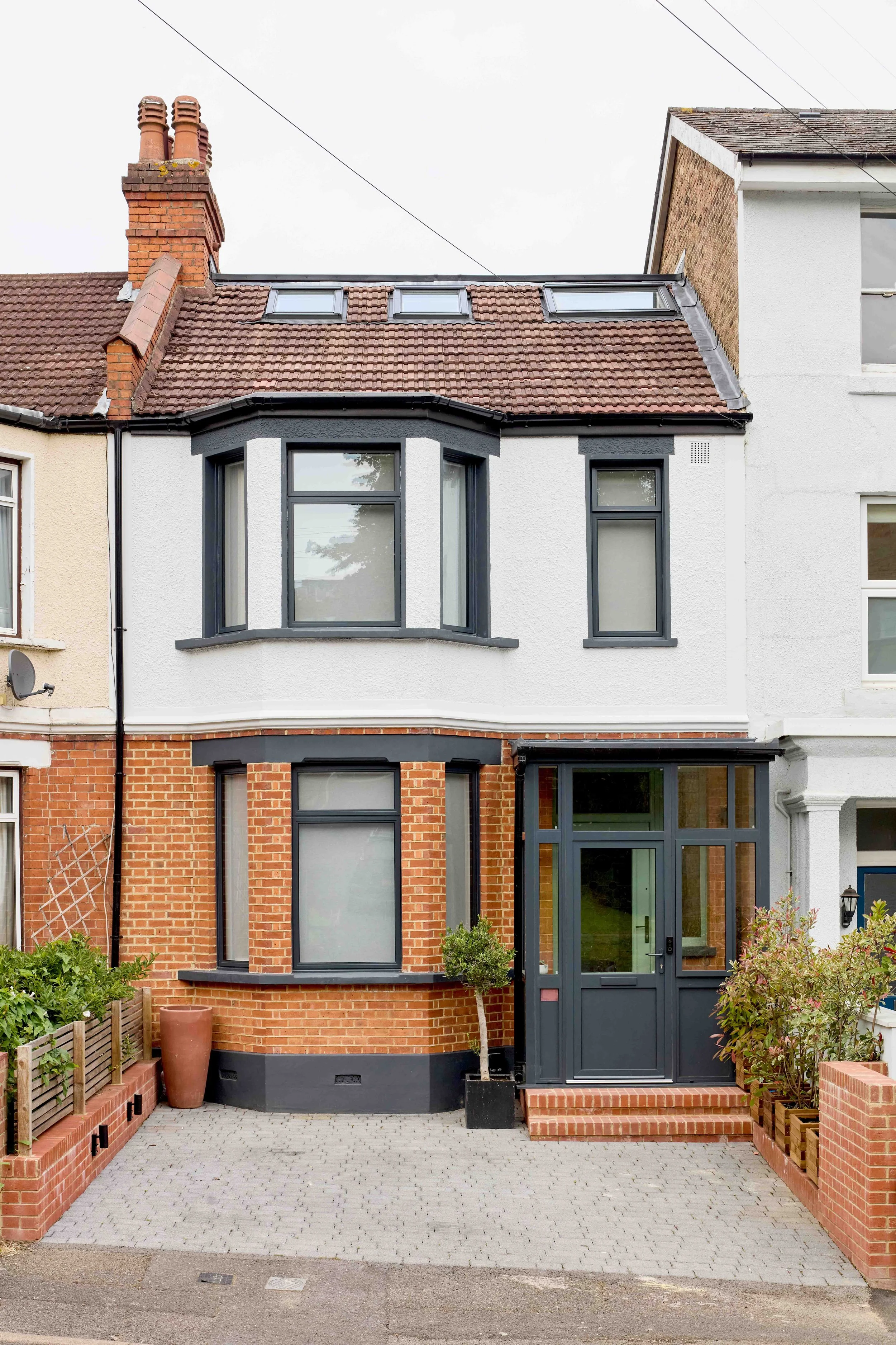 Front view of a two-story house with white upper walls and red brick lower walls. The house features large front windows with black frames, a glass front door, and a small paved pathway. There are plants and a potted tree near the entrance.