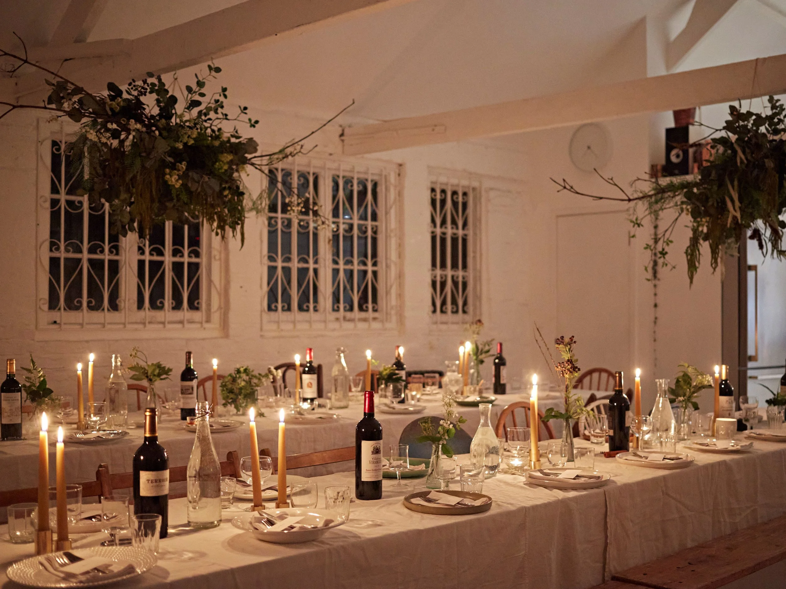 A long dining table set for a dinner event with candles, wine bottles, vases with flowers, plates, glasses, and cutlery in a warmly lit room with three large windows with decorative iron bars.