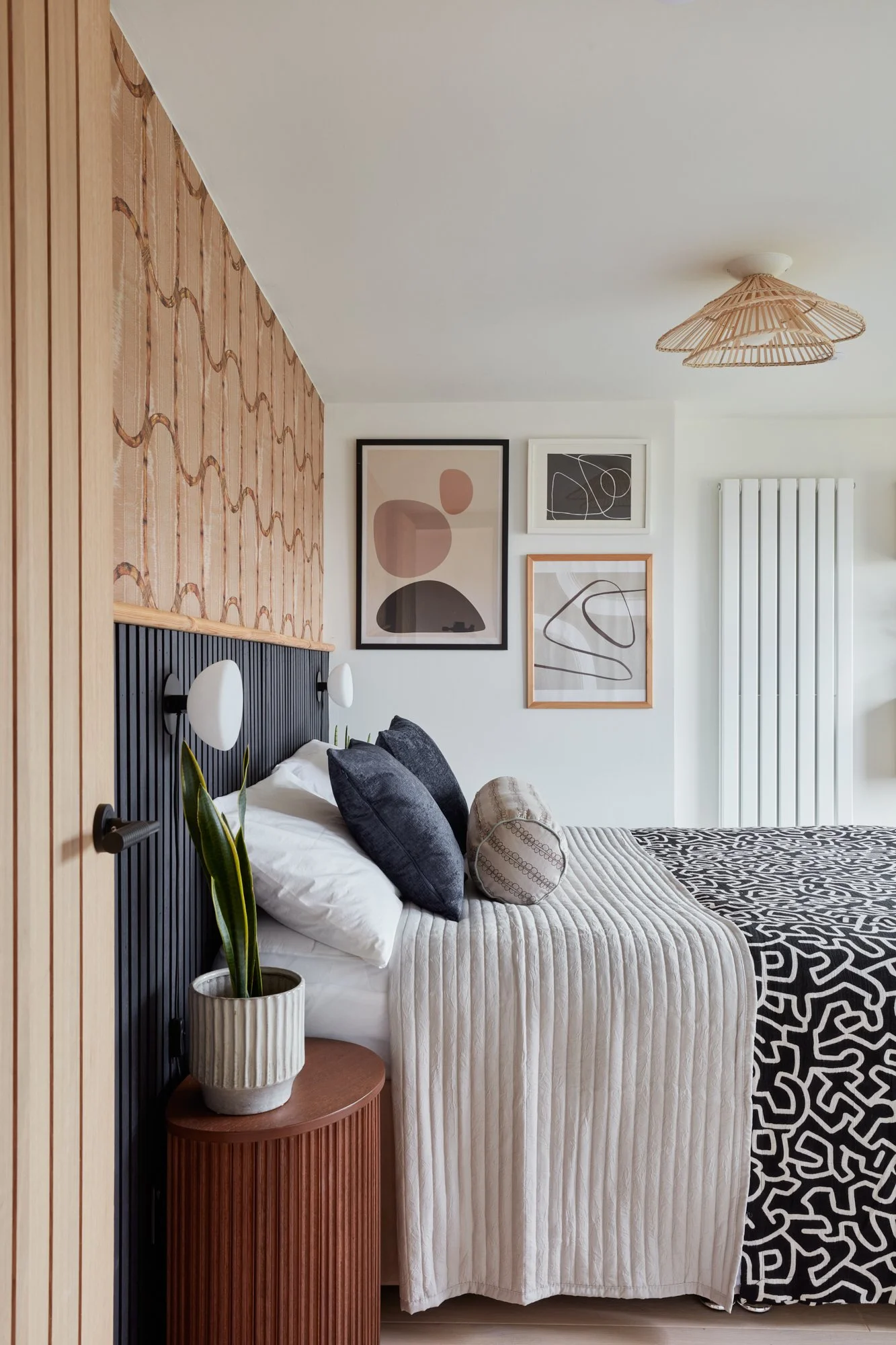 A modern bedroom with a bed featuring black and grey pillows, beige and patterned bedding, and a wooden side table with a potted plant. The wall above the bed has four abstract art pieces. There is a wooden panel and wall lamp on the left, and a whit