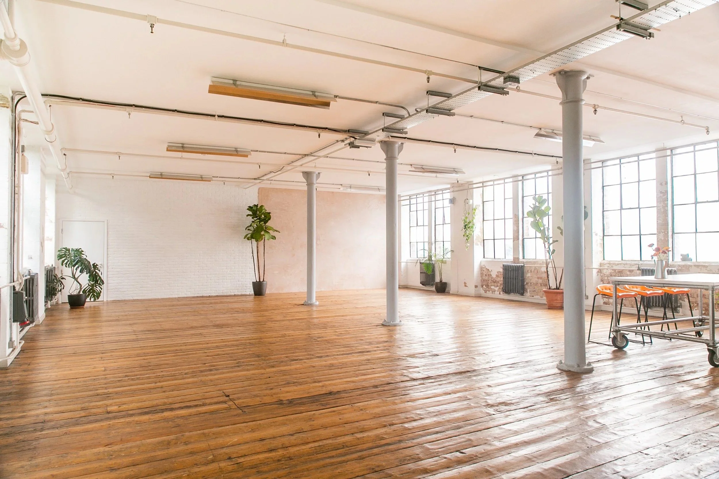 A spacious, bright studio with wooden floors, large windows, and potted plants. The ceiling has exposed pipes and lighting fixtures. There is minimal furniture, including a table with orange chairs and a rolling cart.