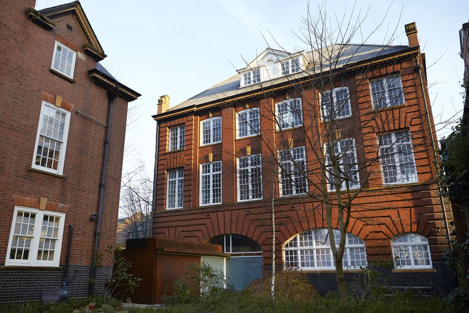 A red brick multi-story building with large white window panes, surrounded by leafless trees and a small grass-covered yard.