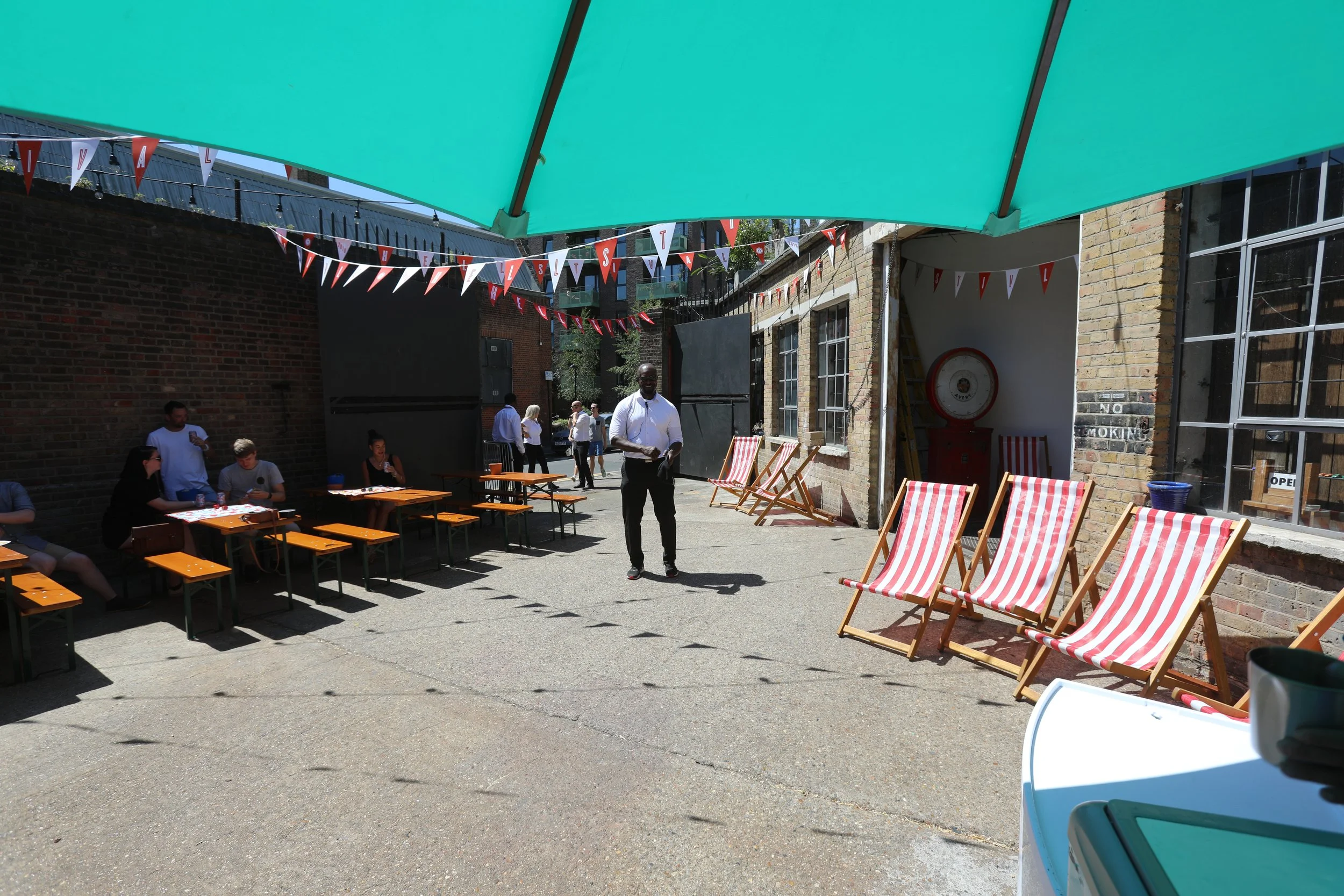 Outdoor patio setup with picnic tables and red-and-white striped deck chairs, under a teal canopy with festive bunting, brick buildings, and several people socializing.
