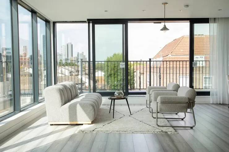 Modern living room with large glass windows, light-colored sofa and armchairs, a small coffee table, and city view outside.