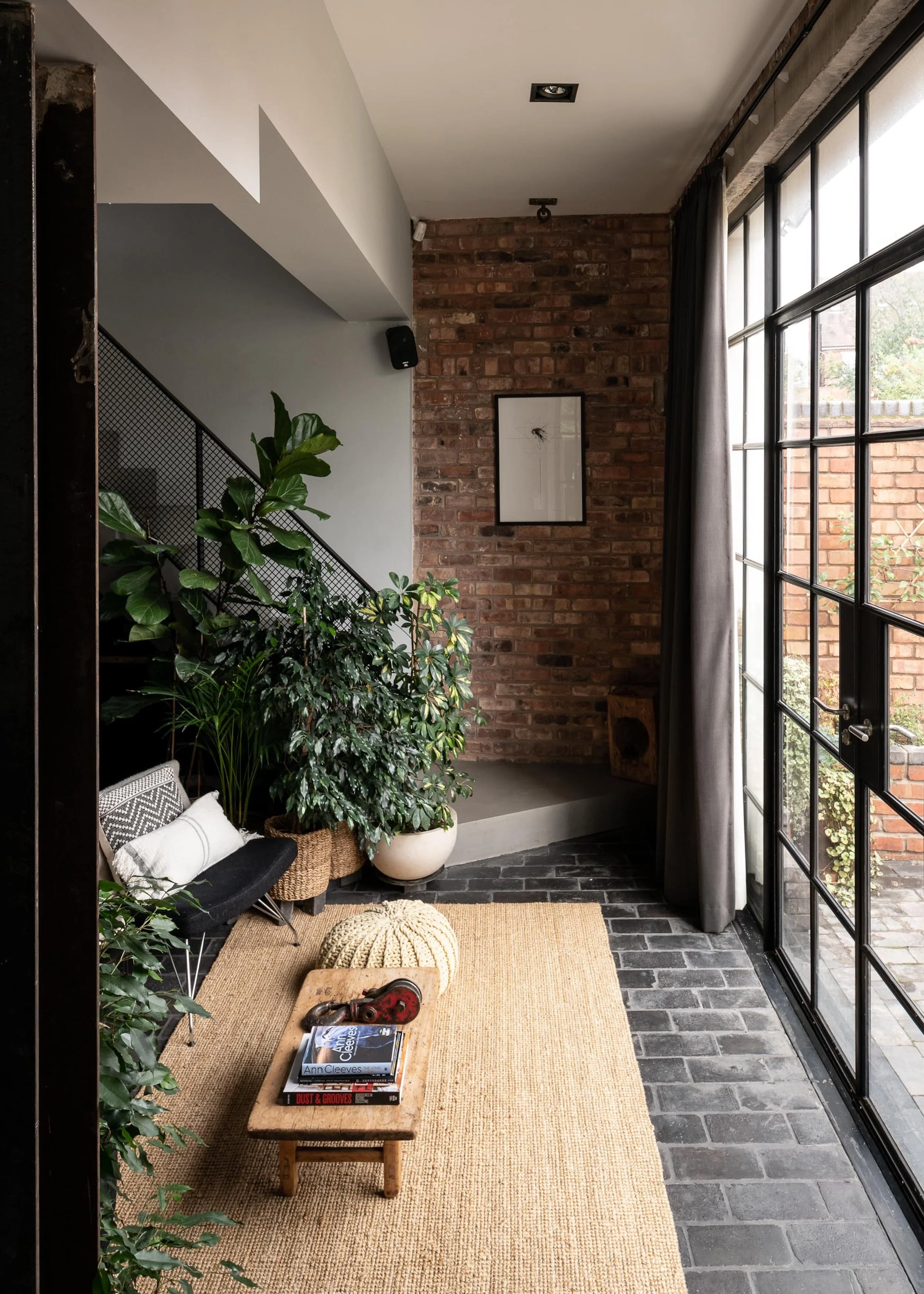 Interior view of a modern living space with large windows, a brick wall, green plants, a small wooden table with books and decorative items, a black bench with cushions, a beige rug, and minimalistic decor.