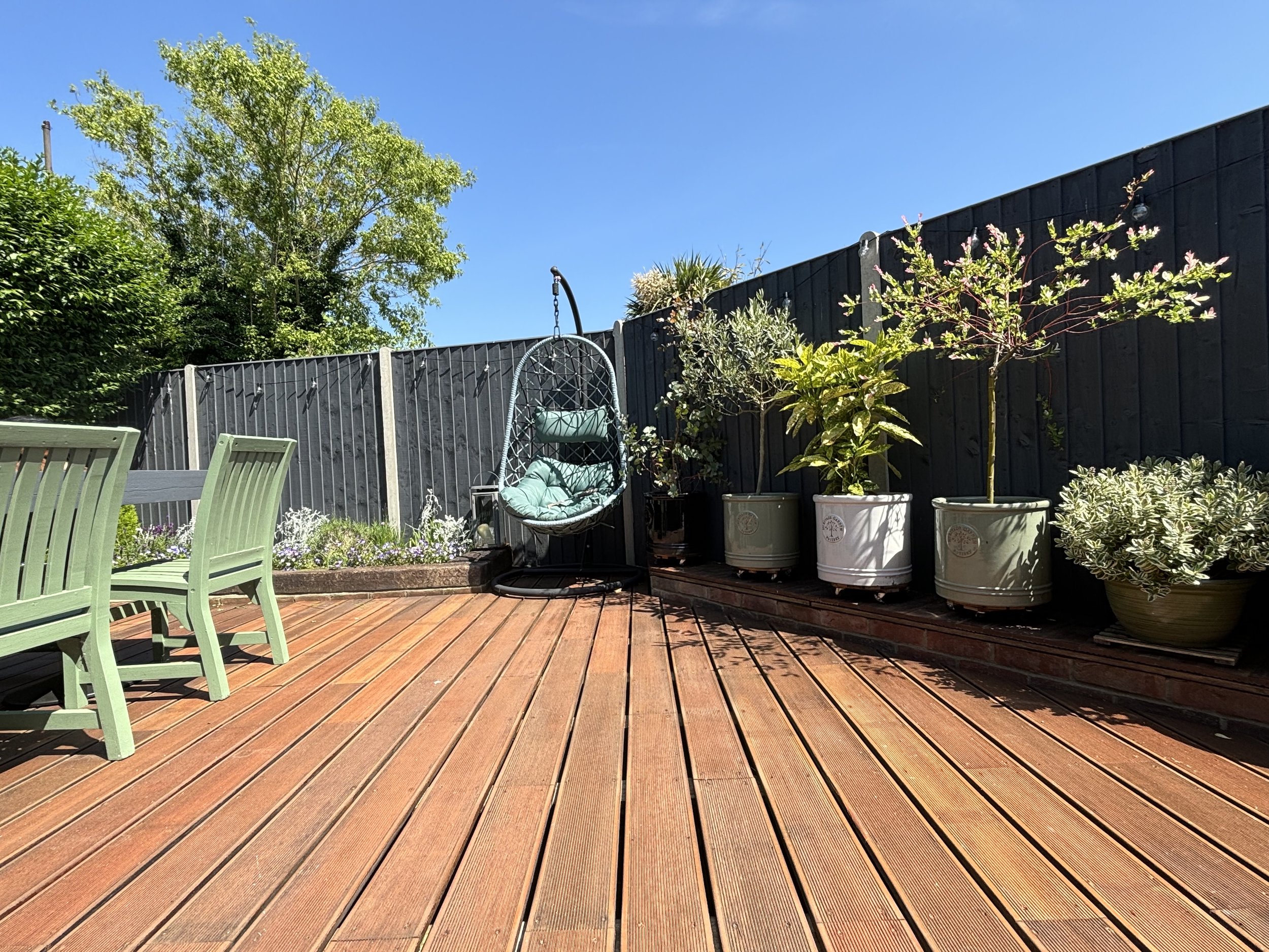 A wooden deck with green chairs, a hanging egg-shaped chair, and potted plants along a black fence on a sunny day with a clear blue sky.