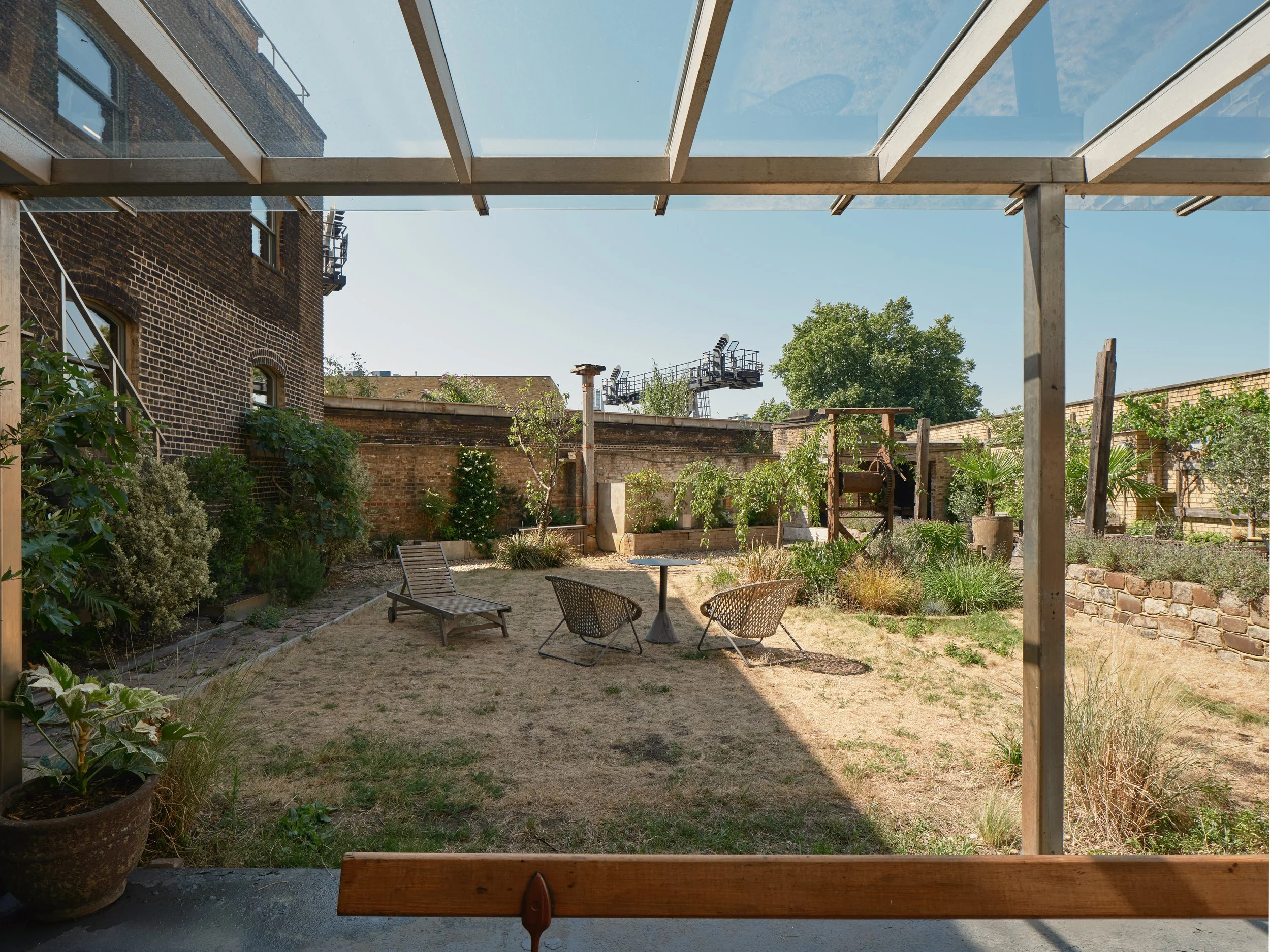View of a backyard garden through a glass roof, with a small table and chairs, a lounge chair, plants, and a brick wall.