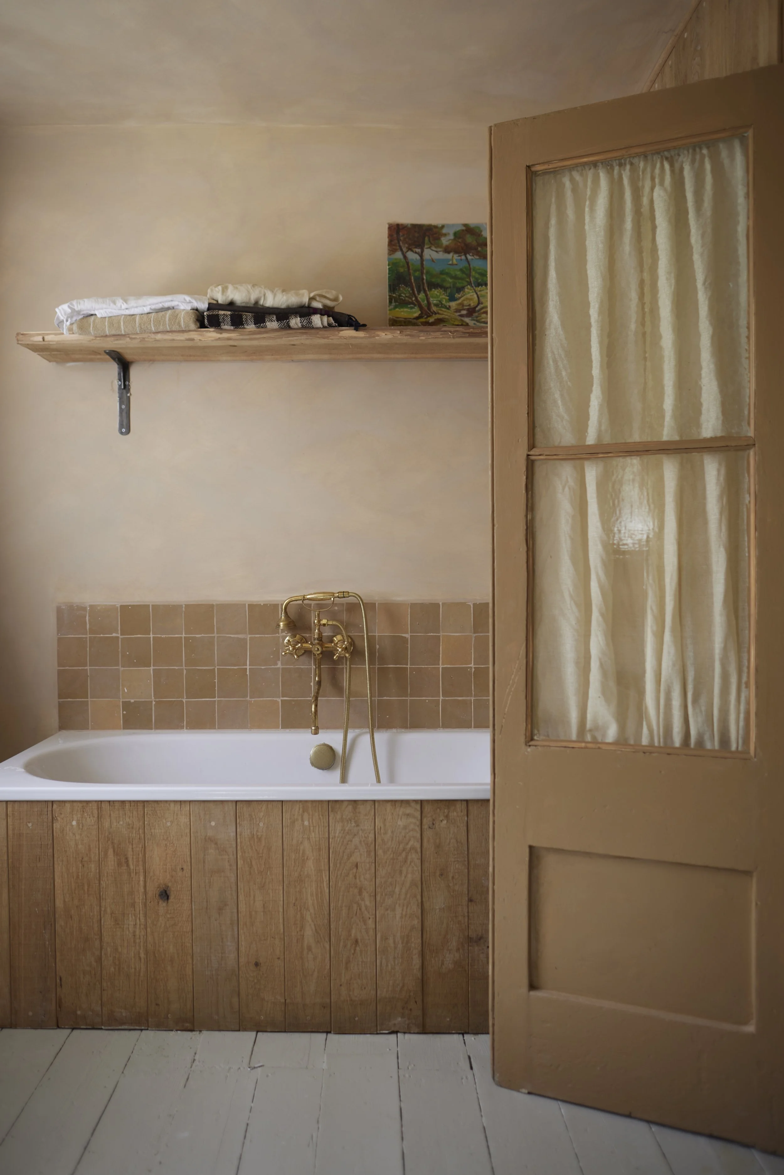 A vintage bathroom with a white bathtub, brass fixtures, wooden paneling, a tiled wall, a wooden door with a frosted glass window, a shelf with folded towels and a landscape painting.