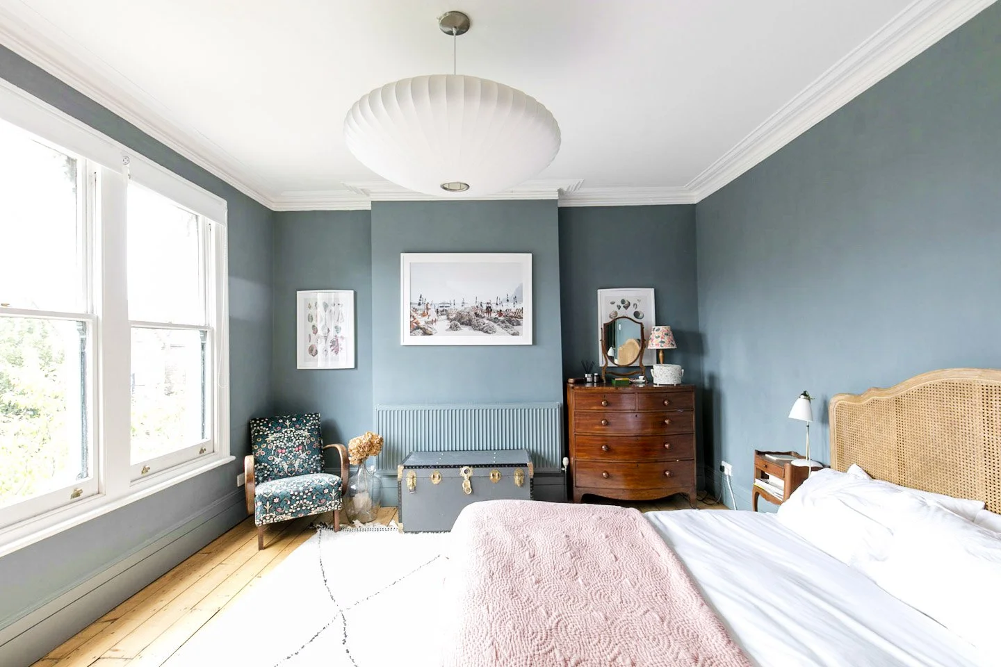 A bedroom with blue walls and white ceiling featuring crown molding. There is a large window on the left, a bed with a rattan headboard and white bedding with a pink bedspread, a wooden nightstand with a lamp, a wooden dresser with a mirror and decor