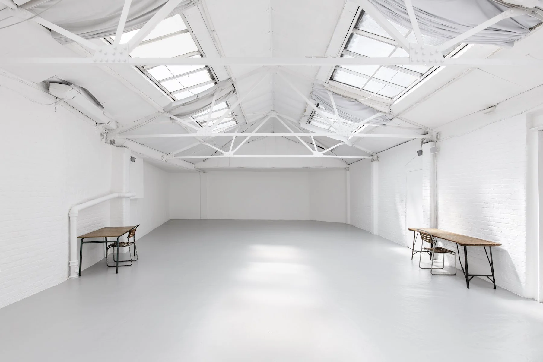 Empty white industrial-style room with skylights, white brick walls, and two small wooden tables with chairs.