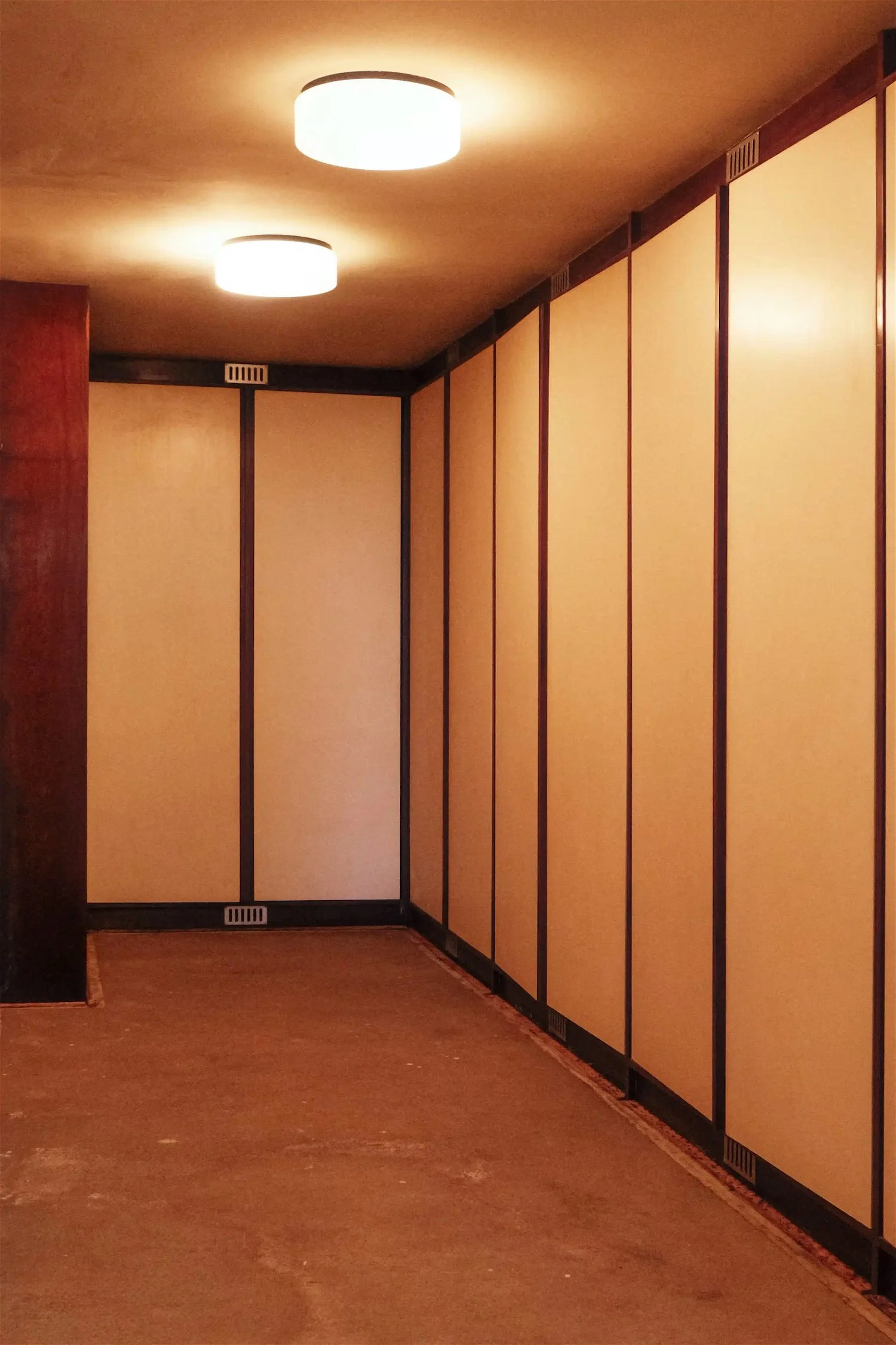 An empty room with beige panel walls, wooden trim and a brown carpet floor. Two large ceiling lights are turned on, illuminating the space.