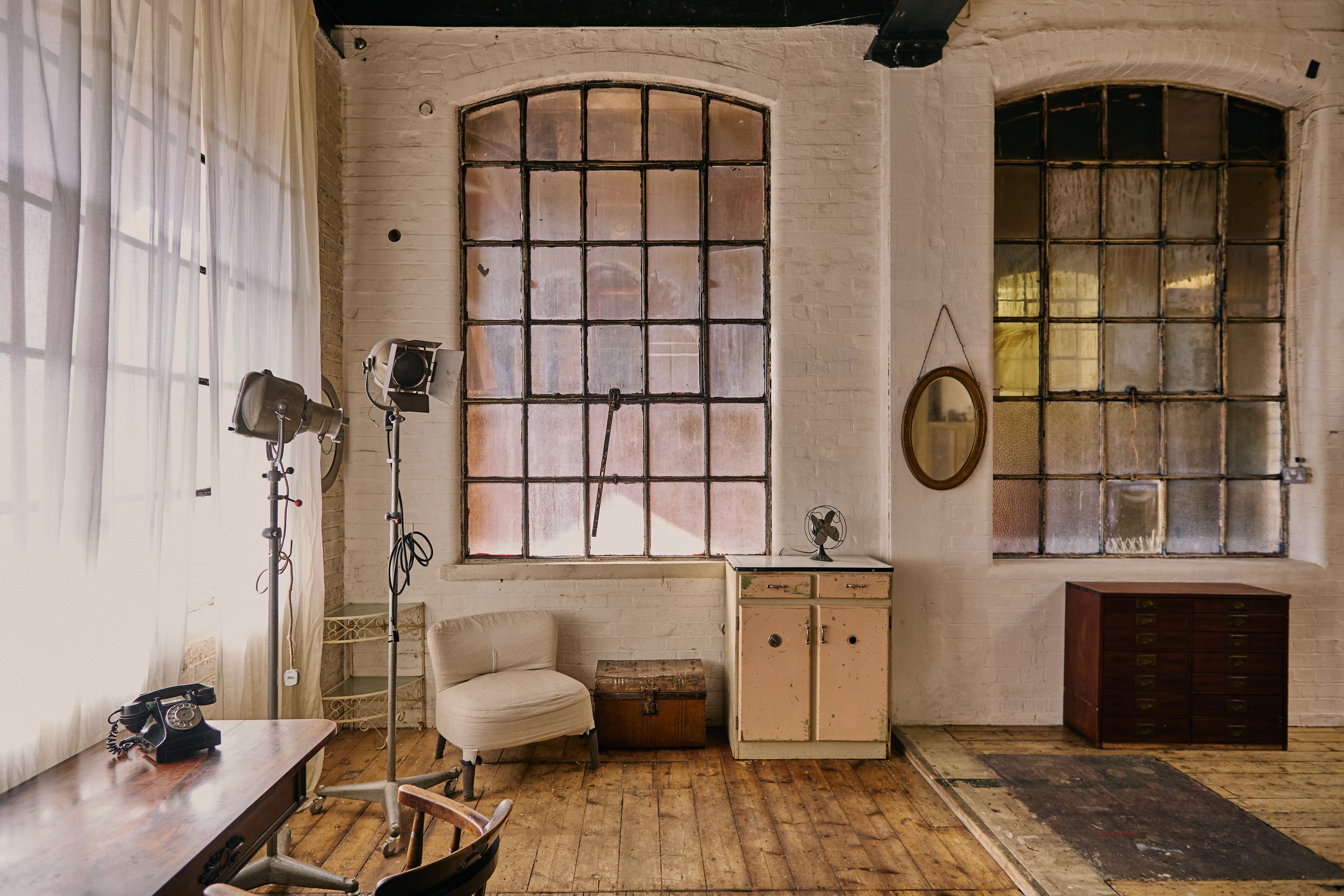 Interior of an industrial-style room with large stained glass windows, white brick walls, and wooden flooring. Furniture includes a vintage desk with a rotary phone, a white upholstered chair, a small cabinet with a fan on top, and a dark wooden ches