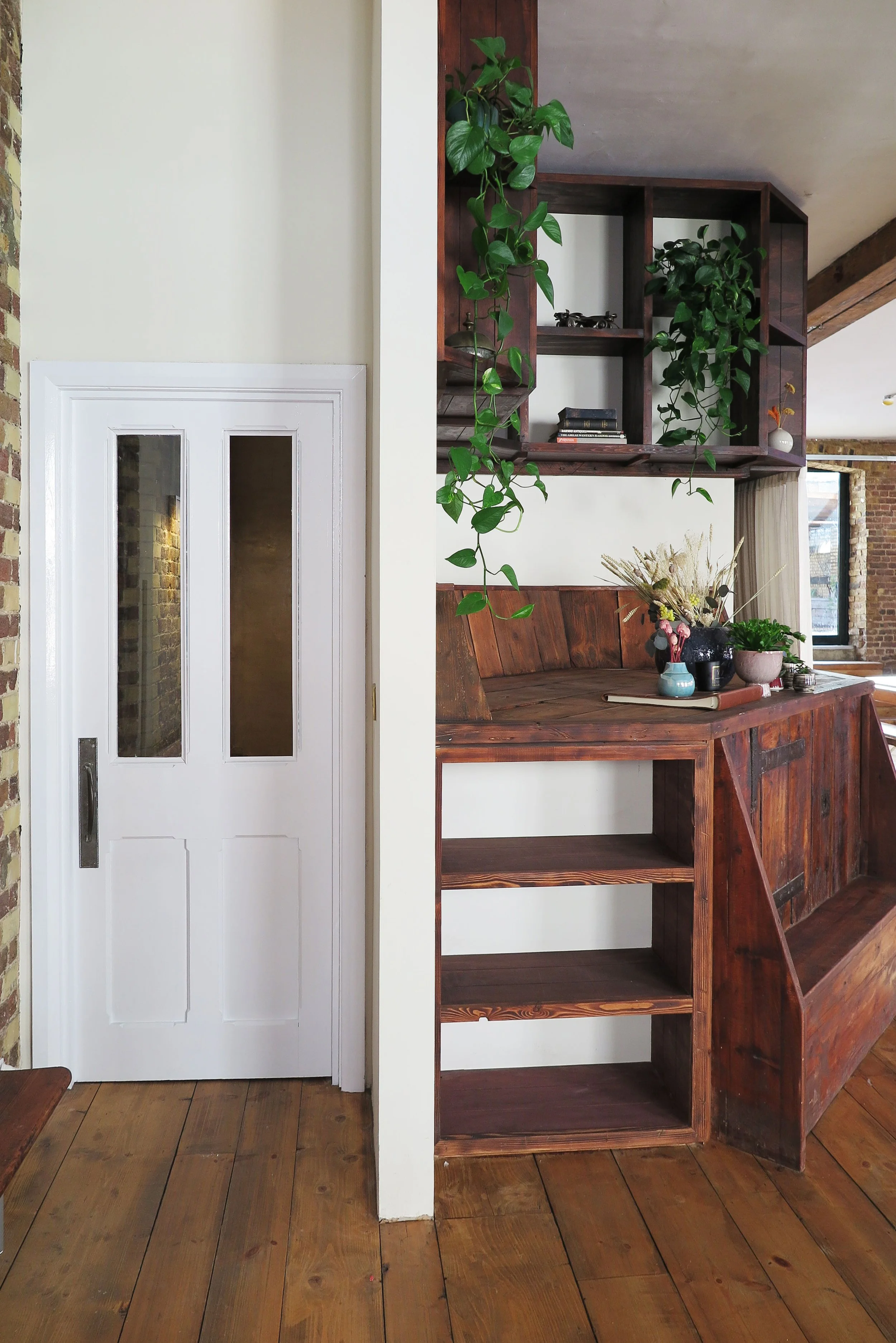 Interior view showing a white door with glass panels, a brick wall, and a wooden shelf with plants and decorative items in a cozy room.
