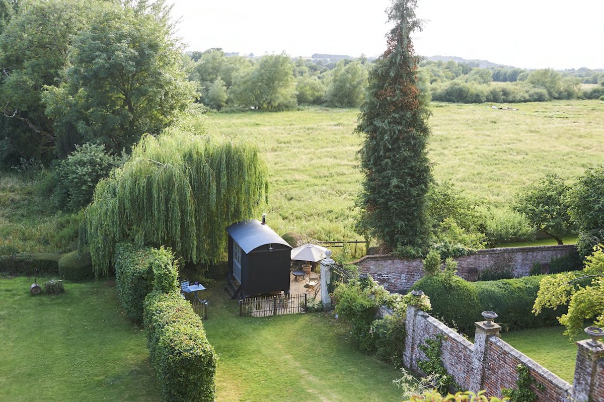 A lush green backyard with a small, black shed, a patio with outdoor furniture, a white umbrella, surrounded by trees and hedges, adjacent to a grassy field and beyond that open countryside.