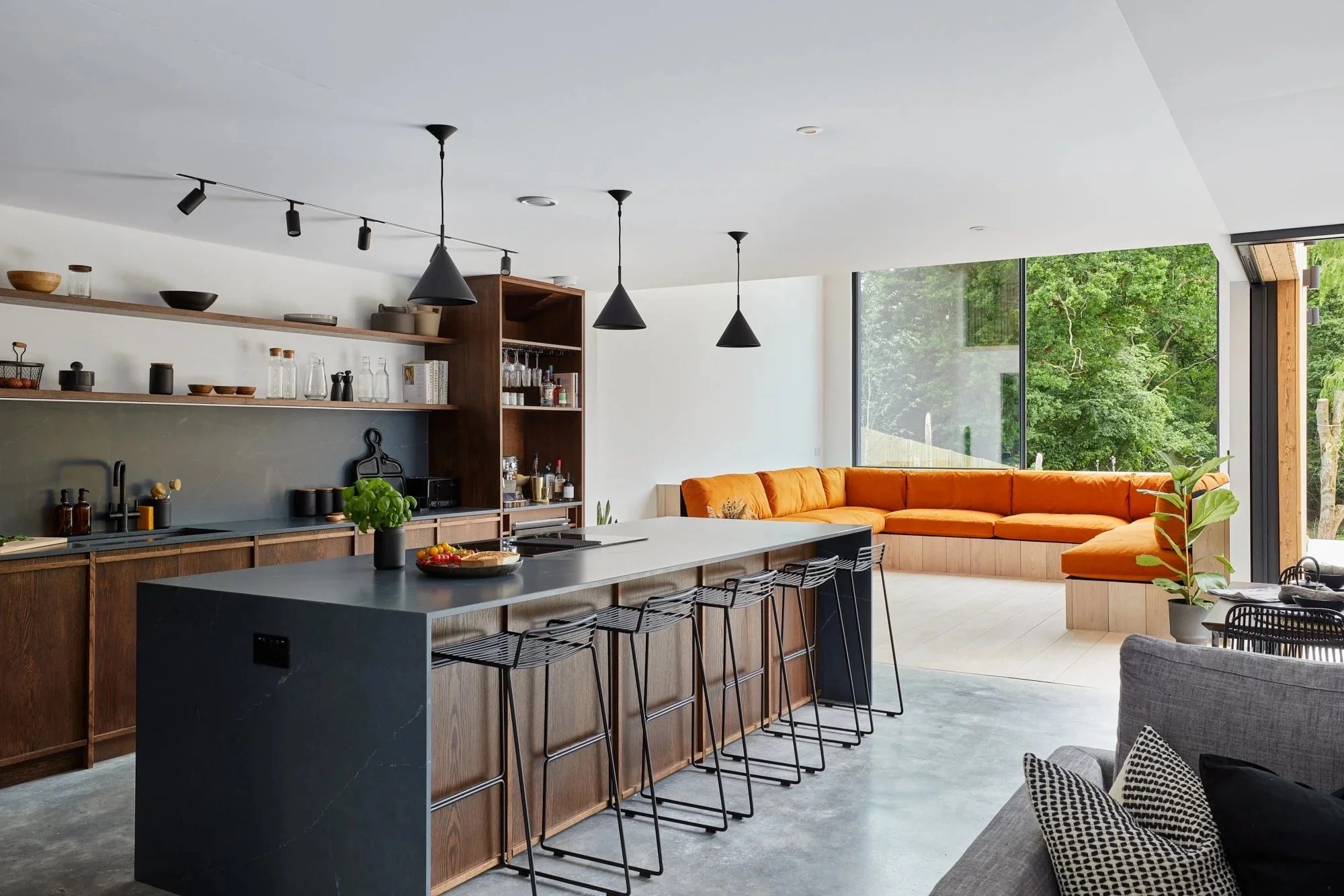Open-concept kitchen and living area with a large black kitchen island, wooden cabinetry, open shelves, and an orange sectional sofa by large windows showing green trees outside.