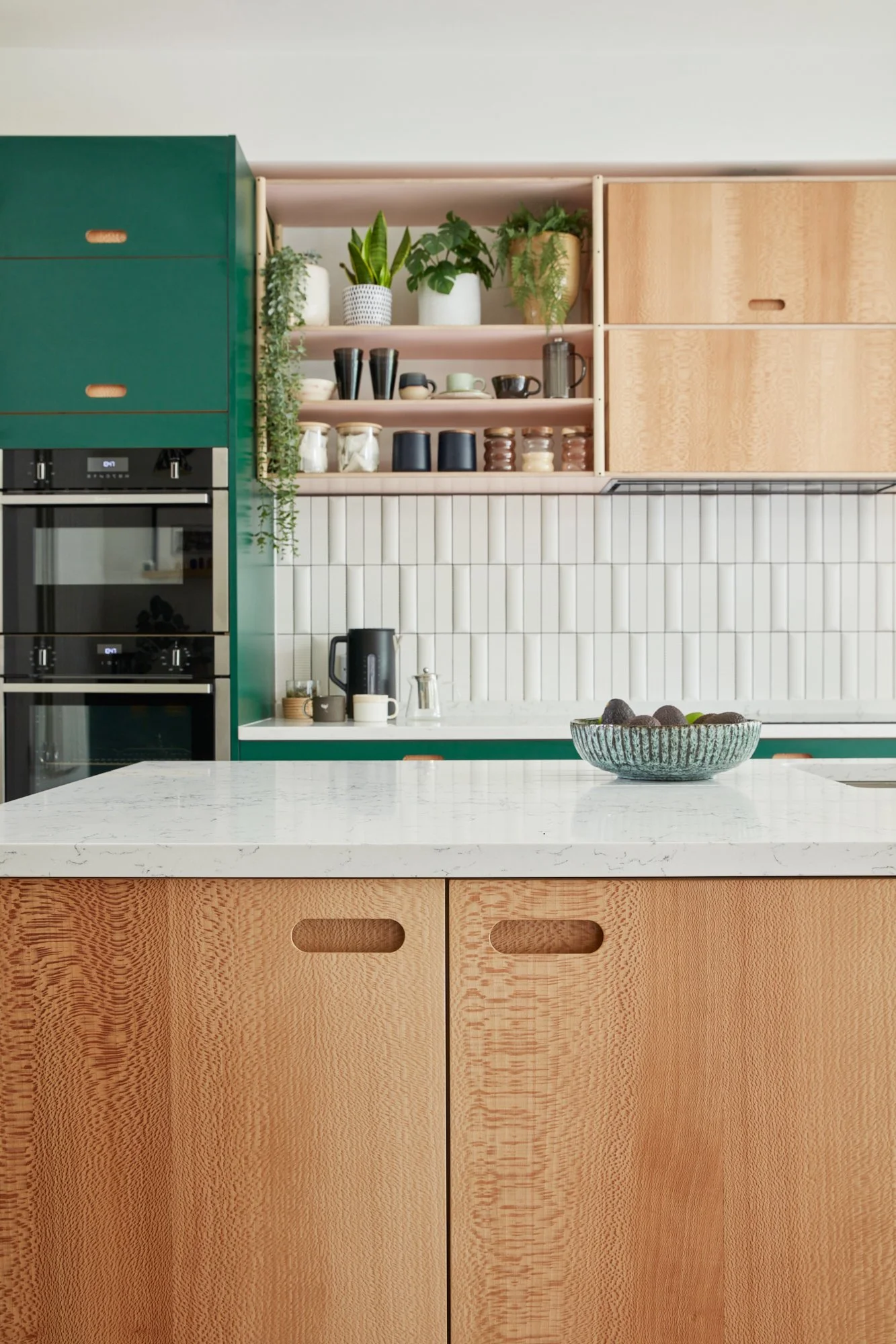 Modern kitchen with a white marble countertop, wooden cabinets, open shelves with plants and dishware, white tiled backsplash, and built-in black oven.